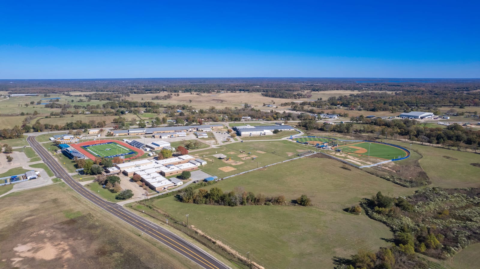 A high aerial over a small town’s schools and sports fields, farmland running to the horizon.