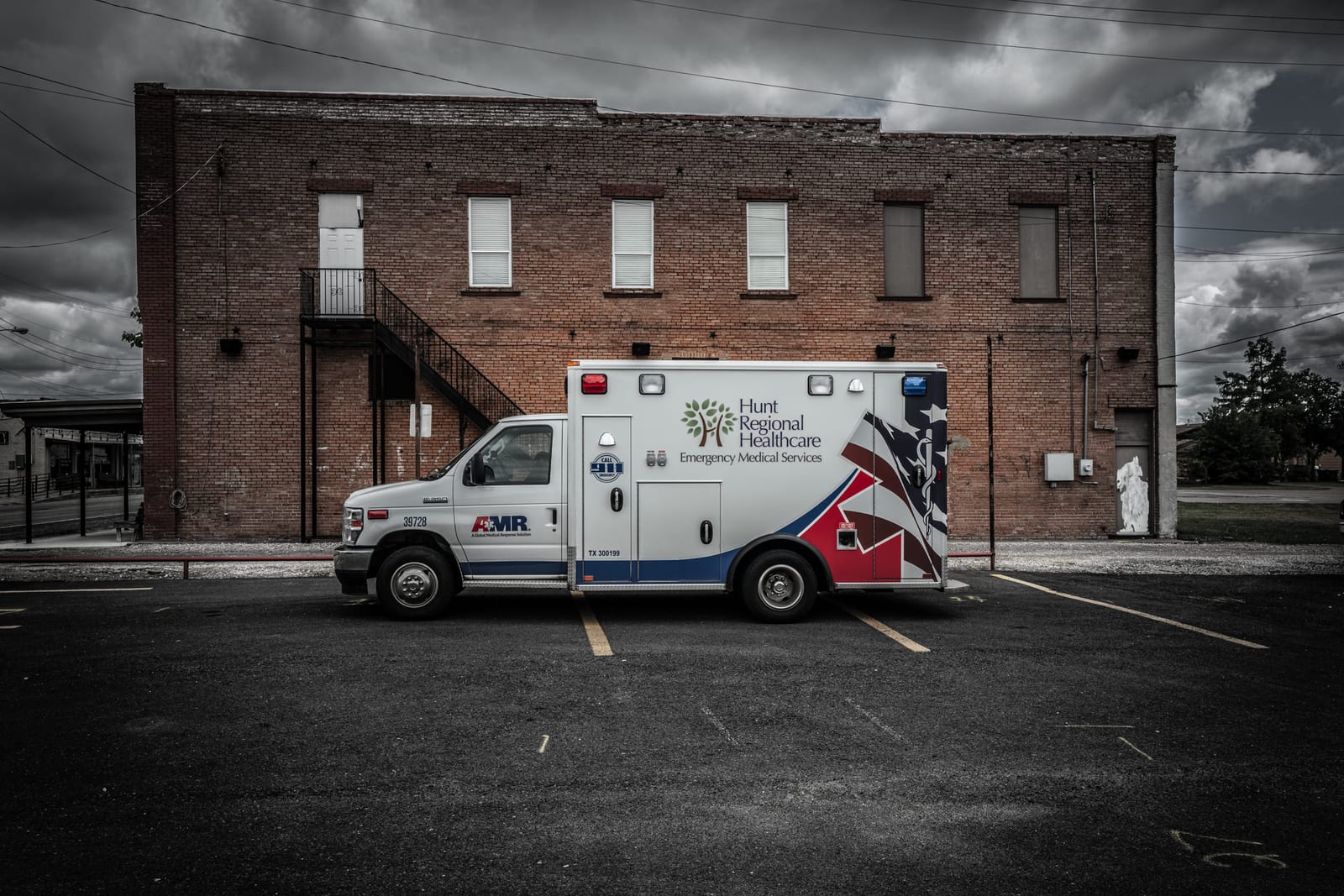 A black-and-white frame of an ambulance parked outside a brick hospital building under heavy sky.