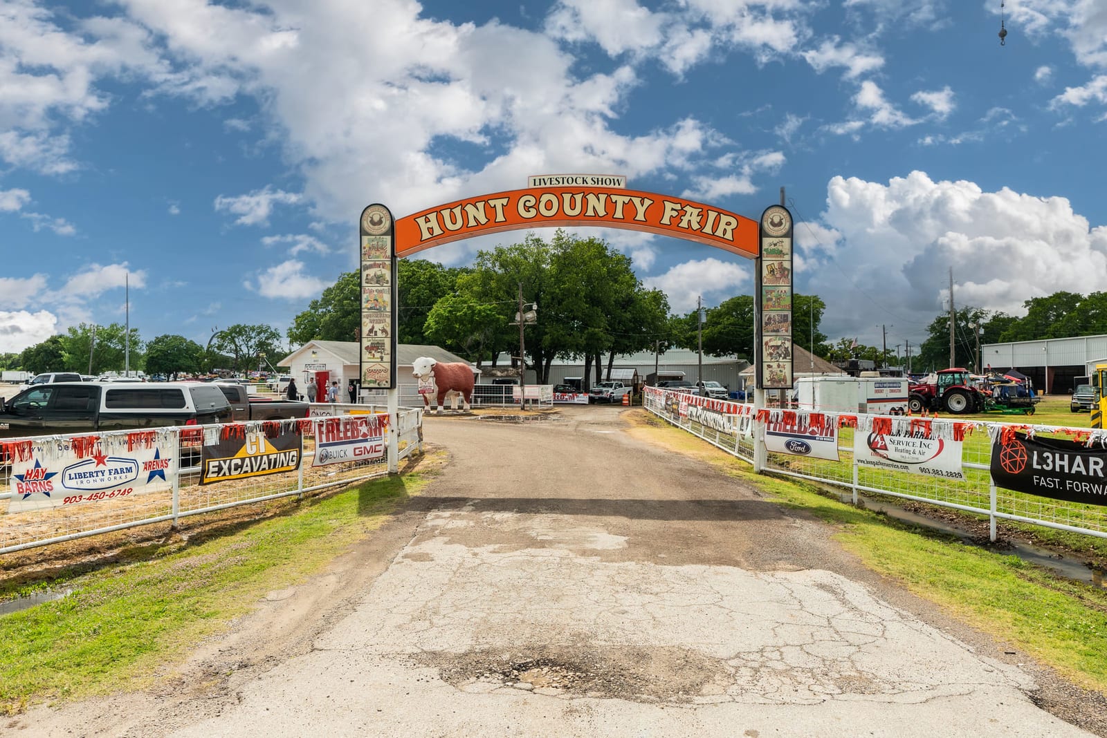 The entrance arch of a county fairground, sponsor banners along both fences.