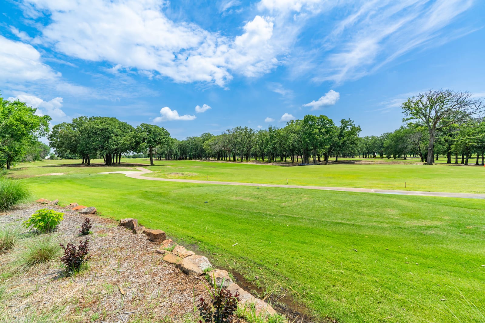 A golf fairway curving between mature trees under a bright, cloud-marked sky.