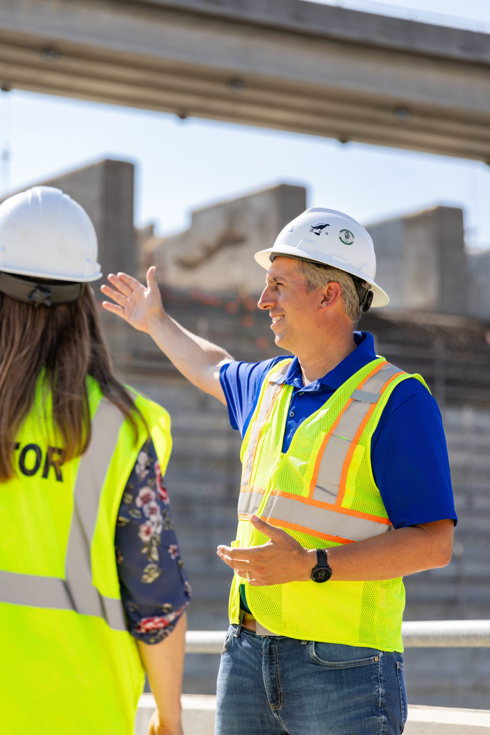 Two workers in hard hats and hi-vis talking on a construction site, one pointing off-frame.