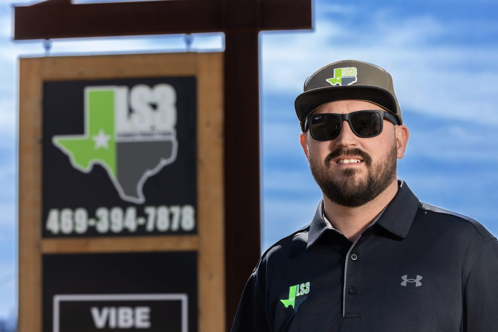 A business owner in a branded cap and polo standing beside his company’s road sign.