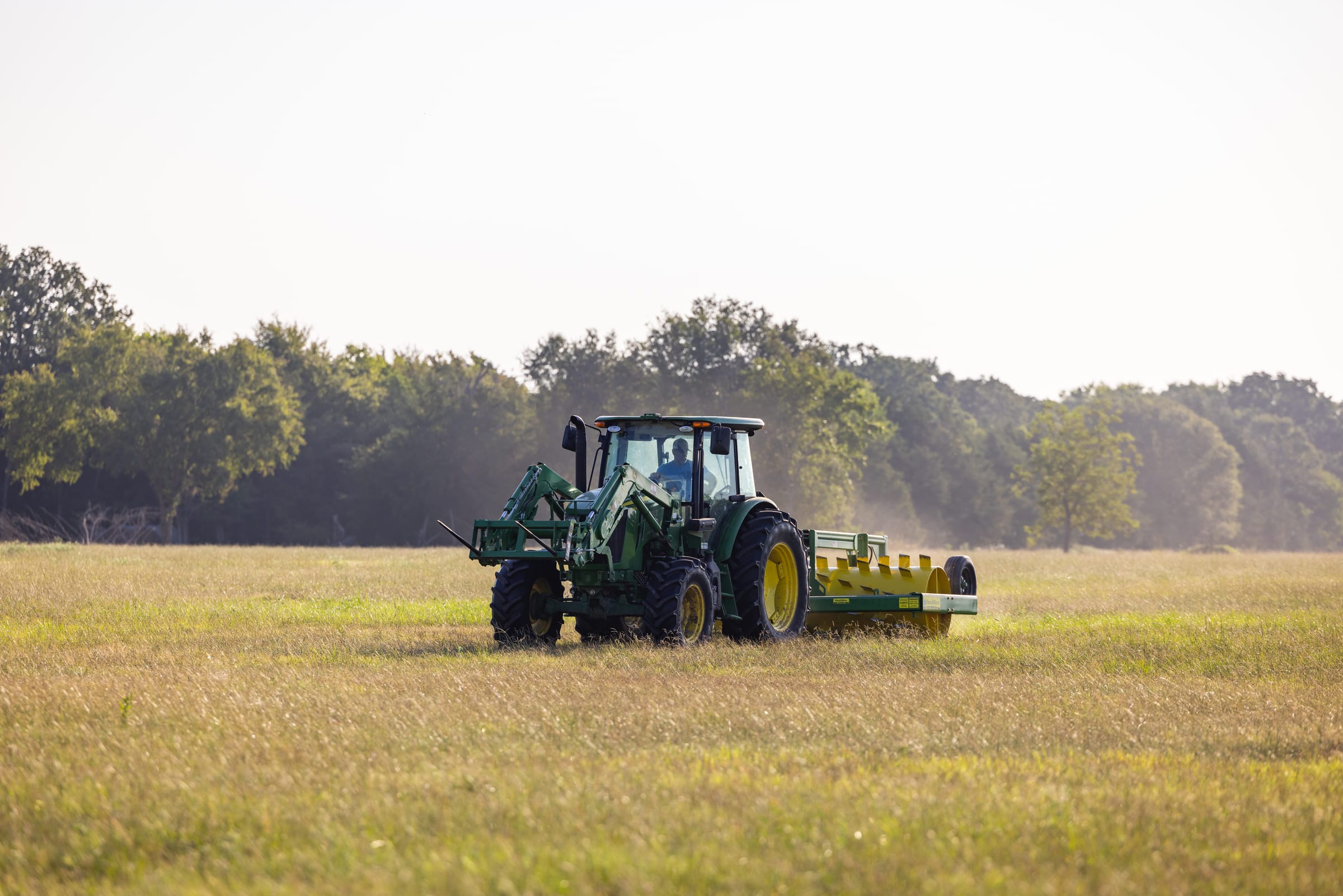 A tractor pulling an aerator across a hayfield at the end of the day, dust lifting behind the implement.