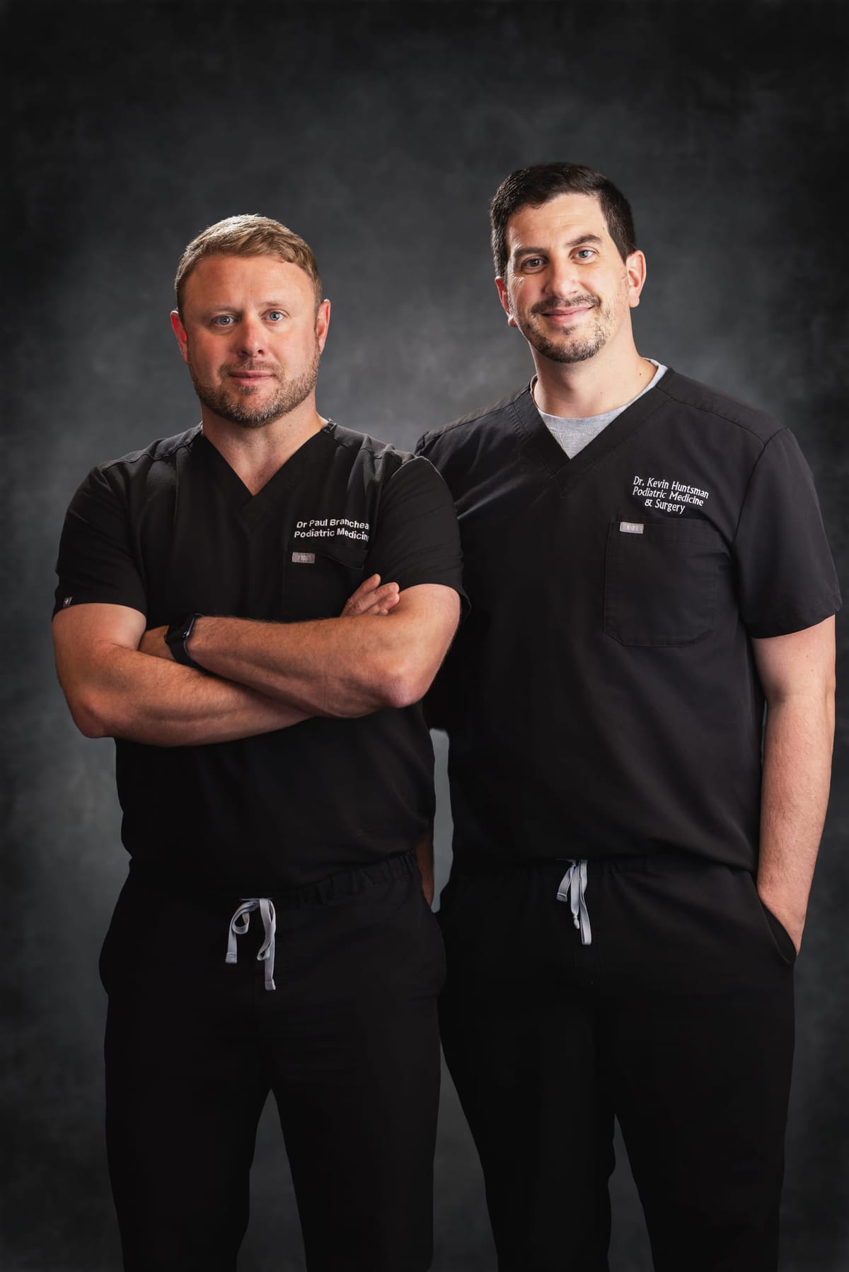 Two practitioners in black scrubs photographed together on a dark studio set.