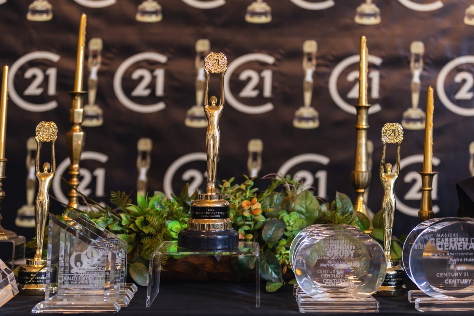 A table of engraved awards and glass trophies dressed with greenery.