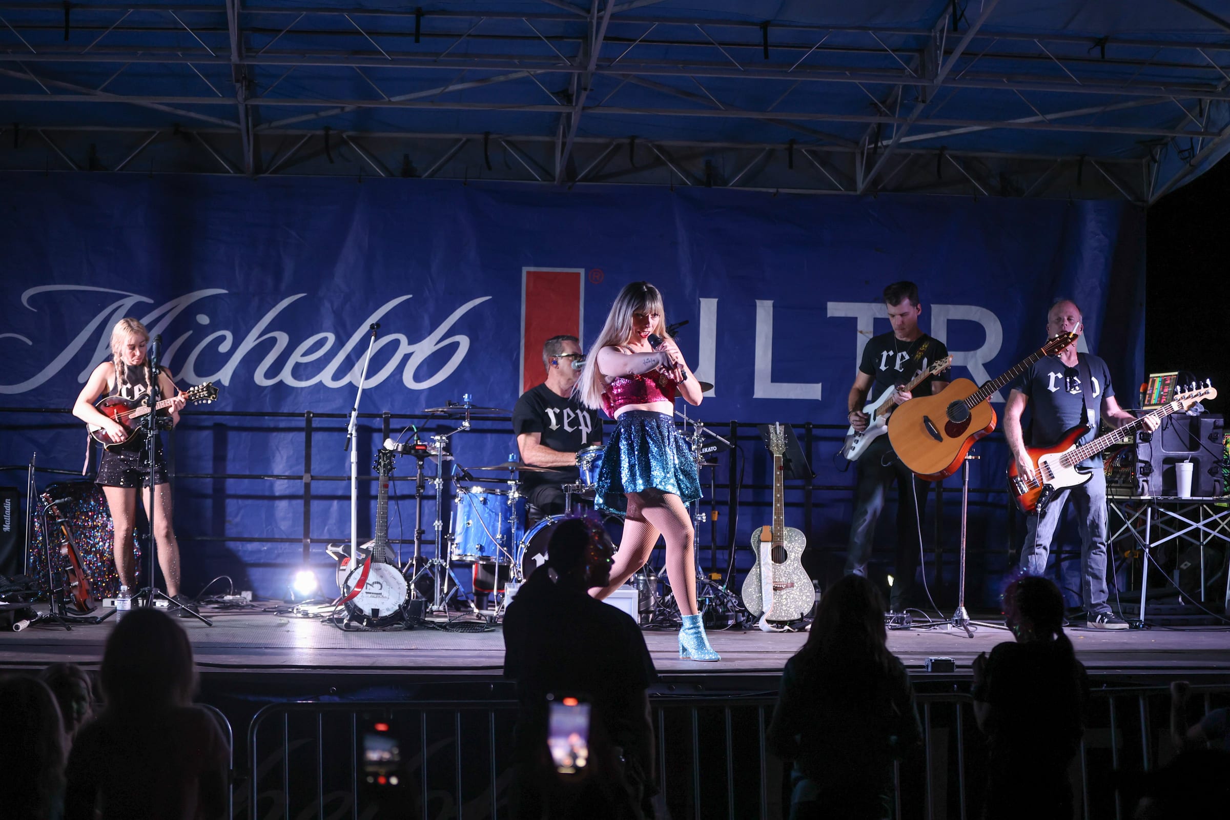 A band on an outdoor stage seen over the heads of the crowd, sponsor banners lit behind them.