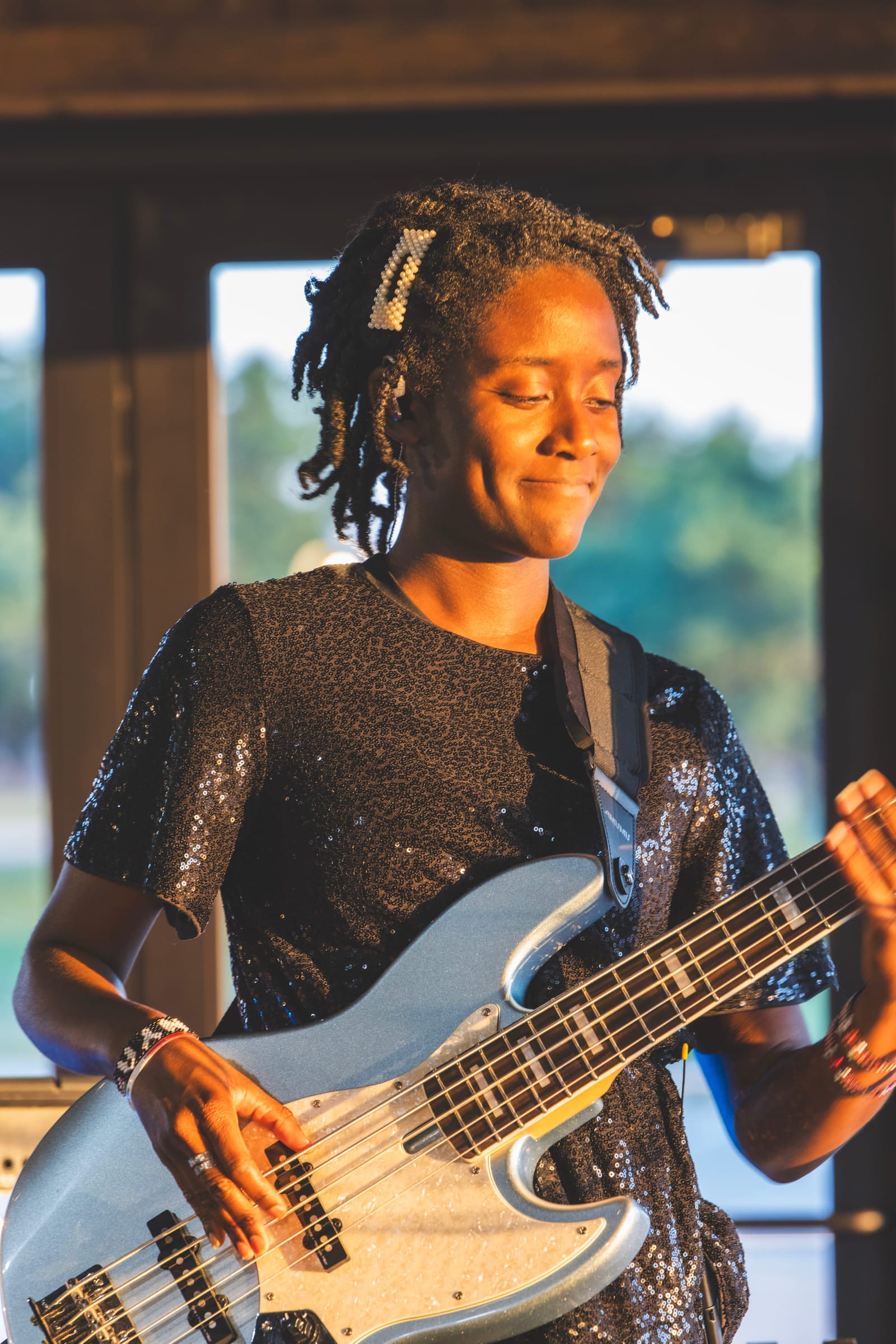 A bass player in a sequinned top mid-phrase, eyes closed, daylight from a doorway behind her.