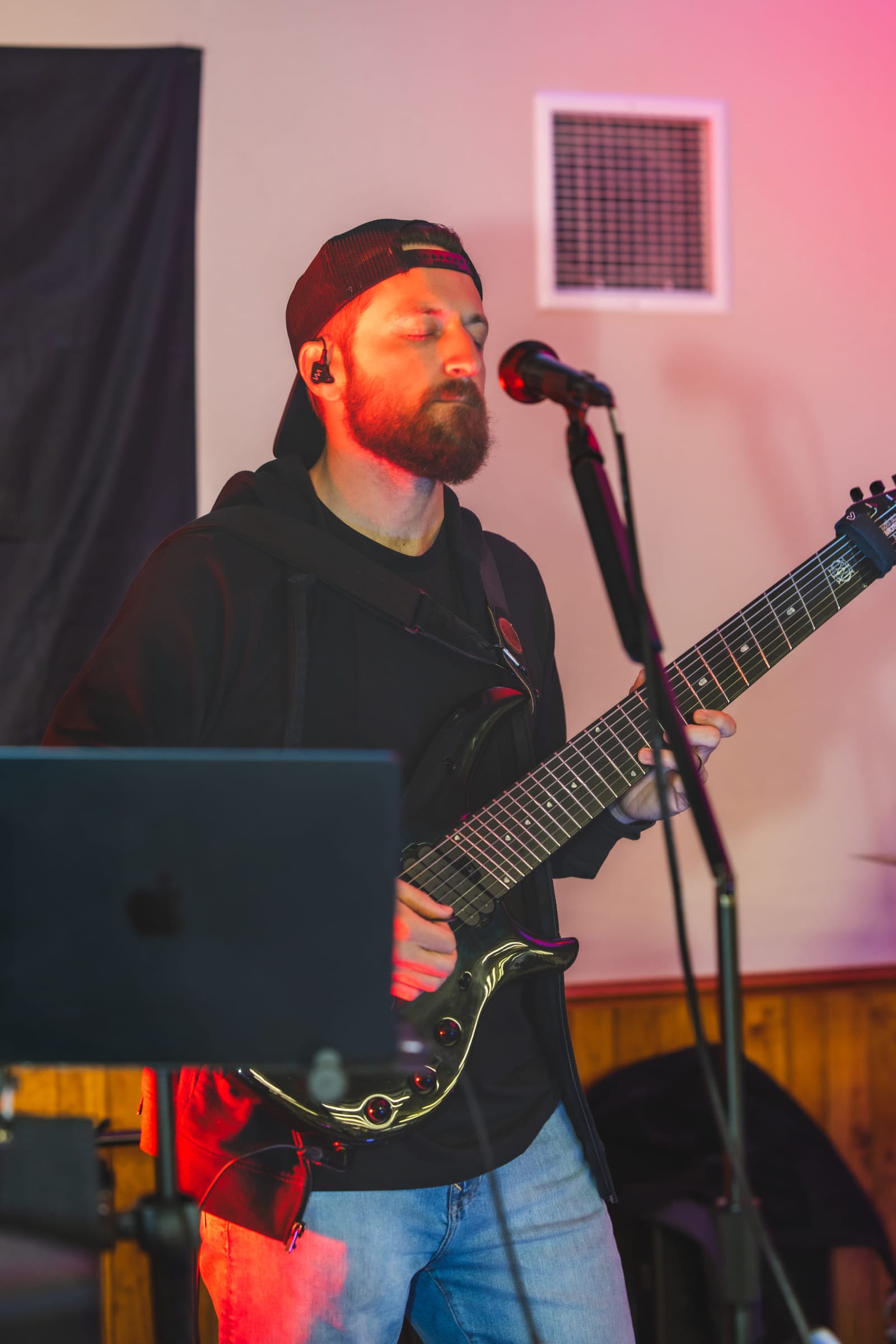 A guitarist singing into the microphone under red and pink stage light.