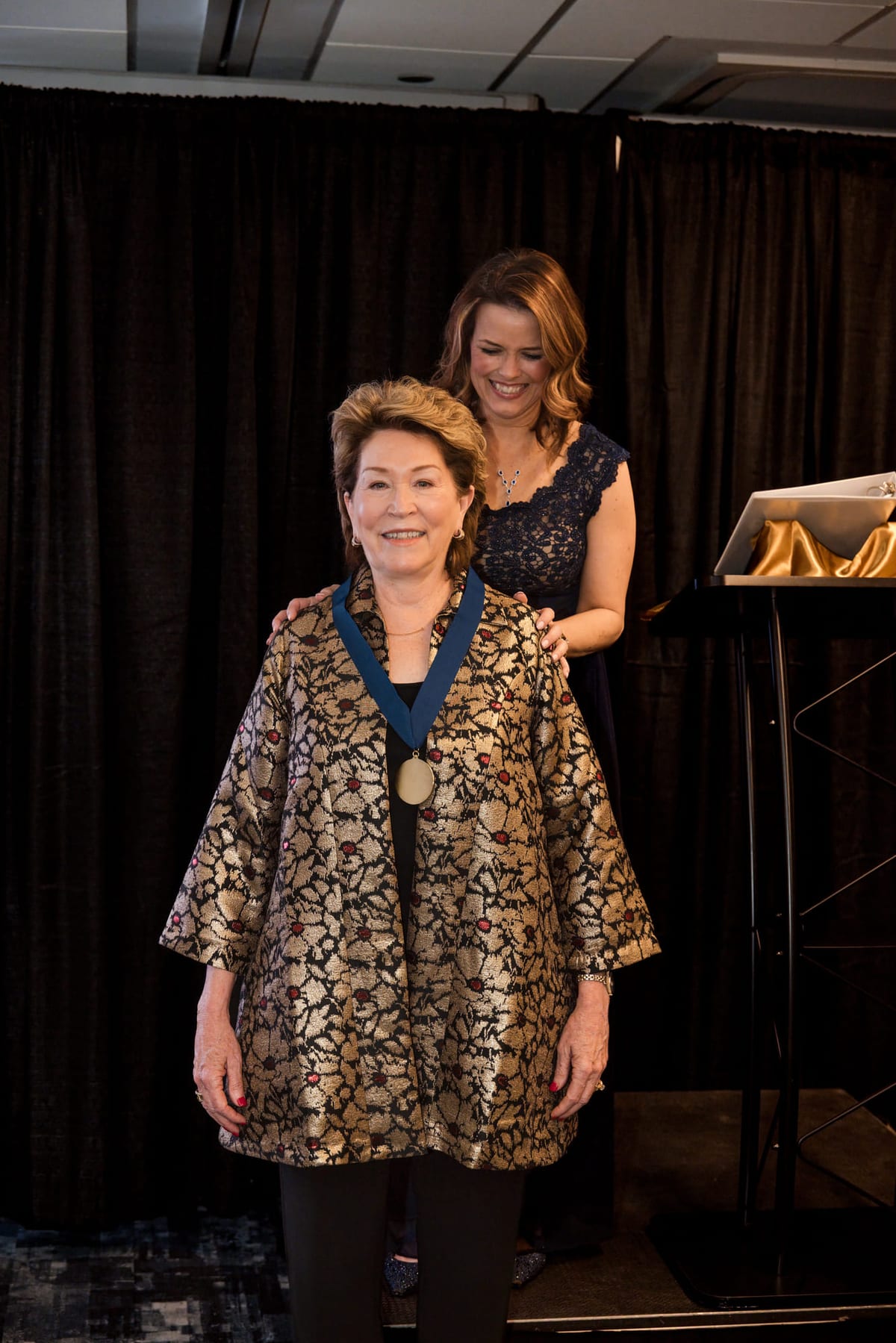 An honoree receiving a medal on stage, a presenter fastening it behind her.