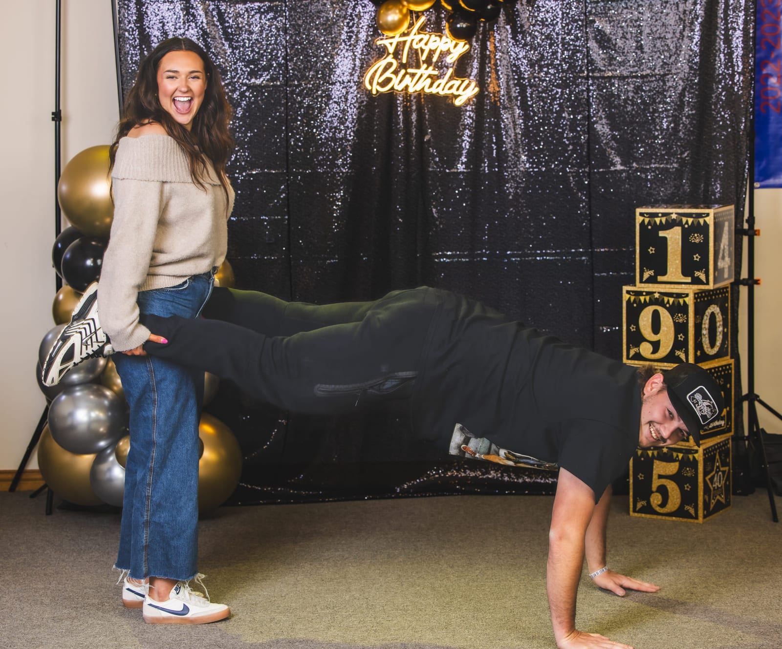 A birthday photo booth, one guest holding a plank while another lifts a leg over him.