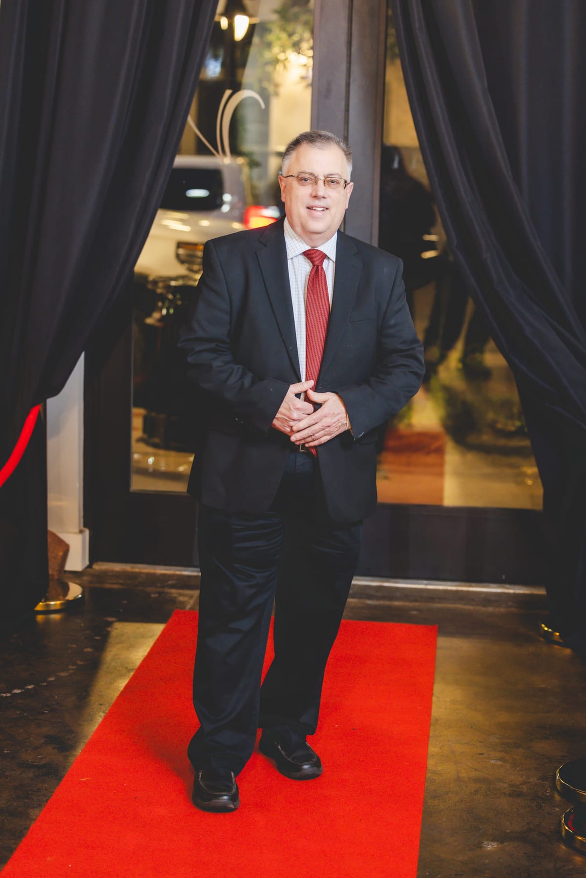 A guest in a dark suit standing on a red carpet between draped curtains.