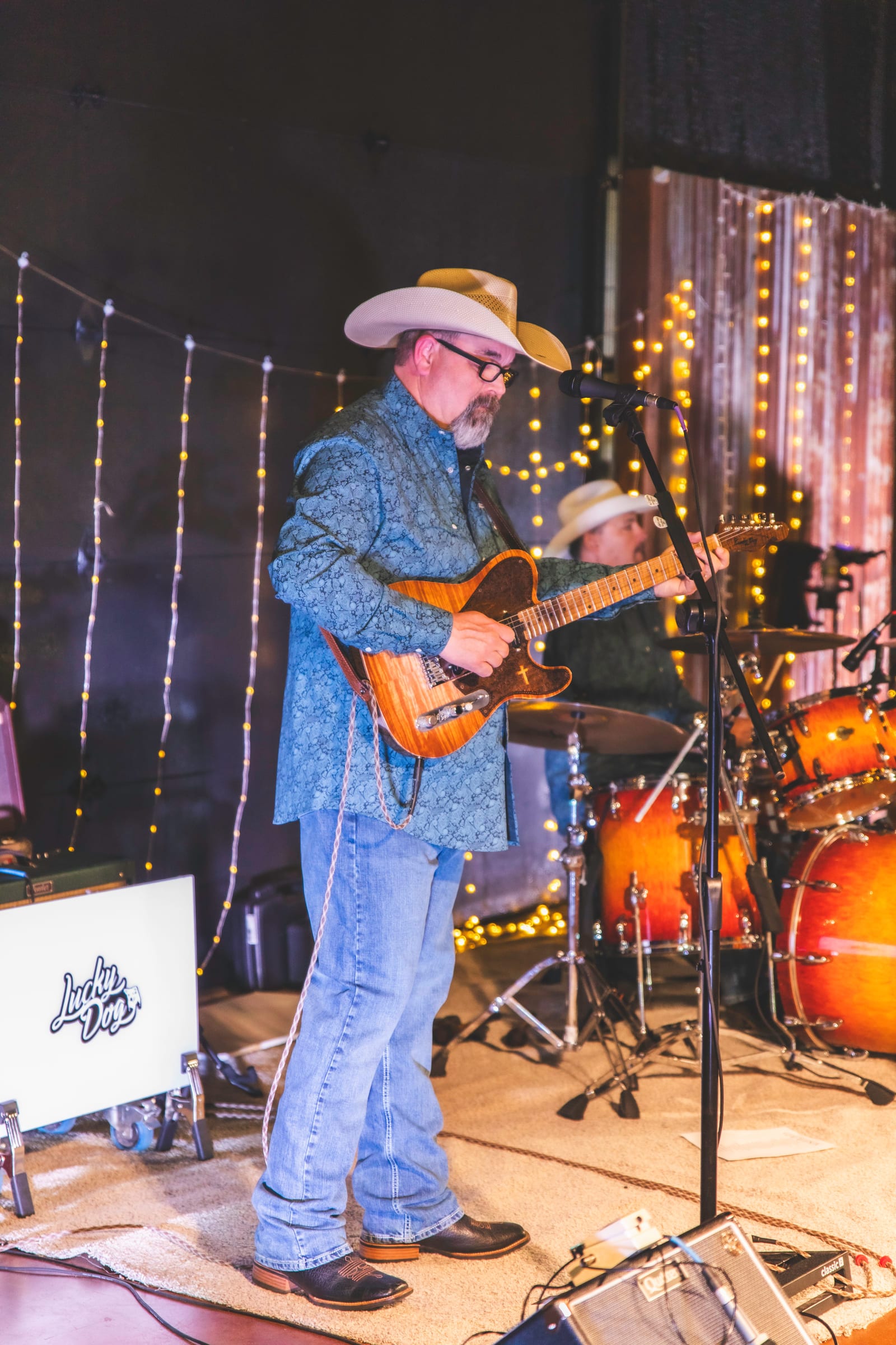 A guitarist in a cowboy hat performing under string lights, a drum kit behind him.