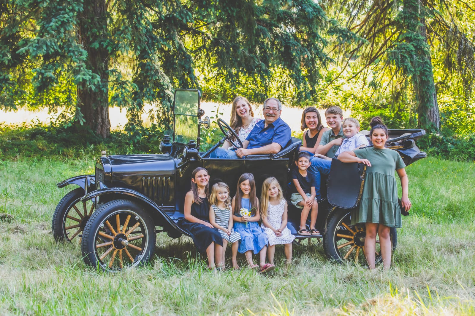 A large family arranged on and around an antique open-top car in a field.