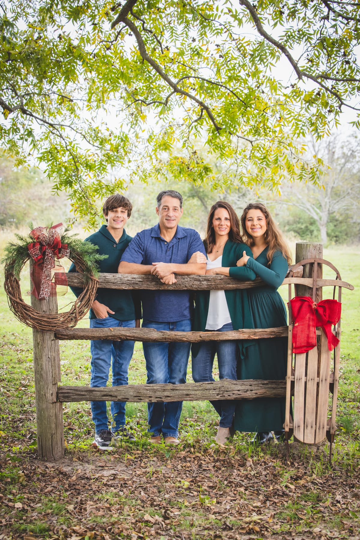 A family of four at a rustic rail bench in the woods, a wreath and a sled propped beside them.
