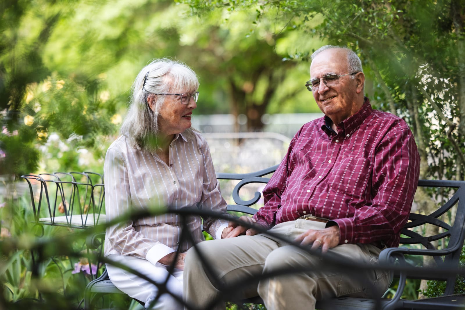 An older couple laughing together on a garden bench under green trees.