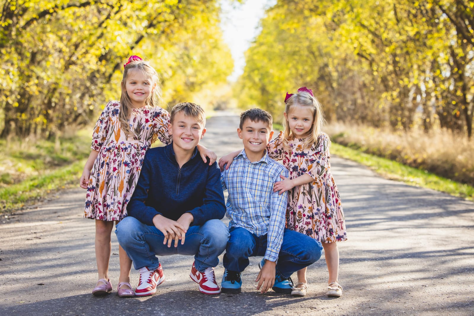 Four children crouched together on a gravel country road lined with trees.