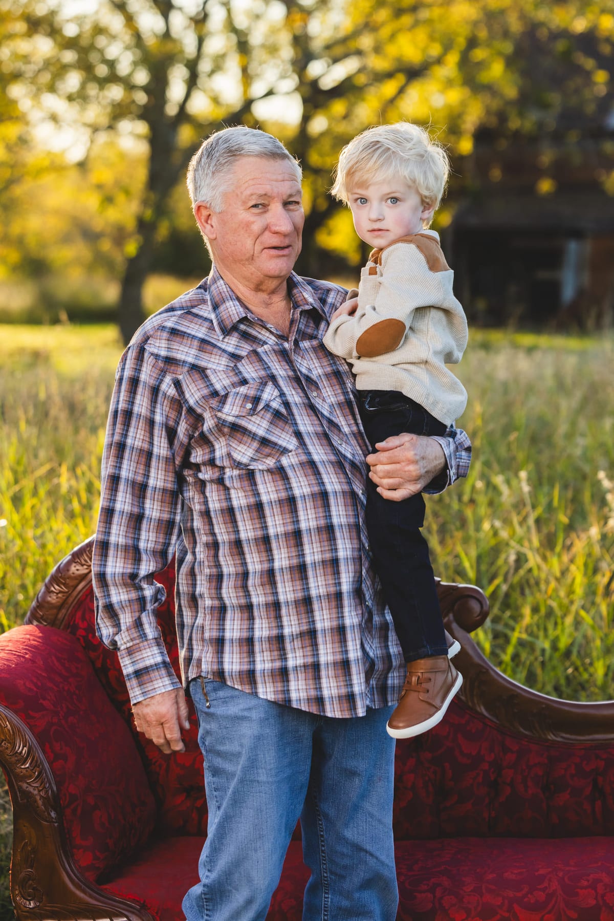 A grandfather holding a small boy beside a red velvet settee set out in a field.