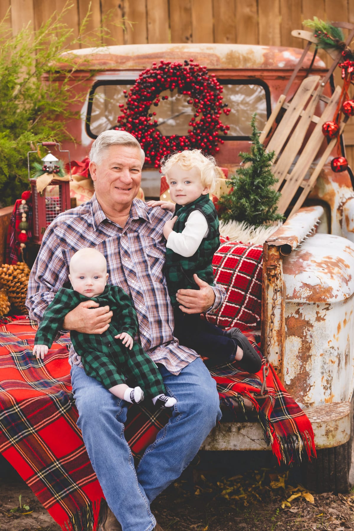 A grandfather on the tailgate of a rusted red truck with two grandchildren, a wreath hung behind them.