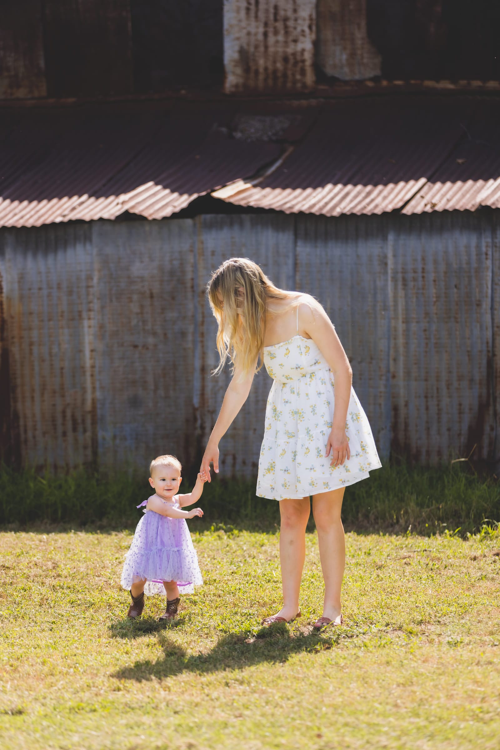 A young woman bending to steady a toddler taking a few steps on the grass in front of a tin-sided barn.