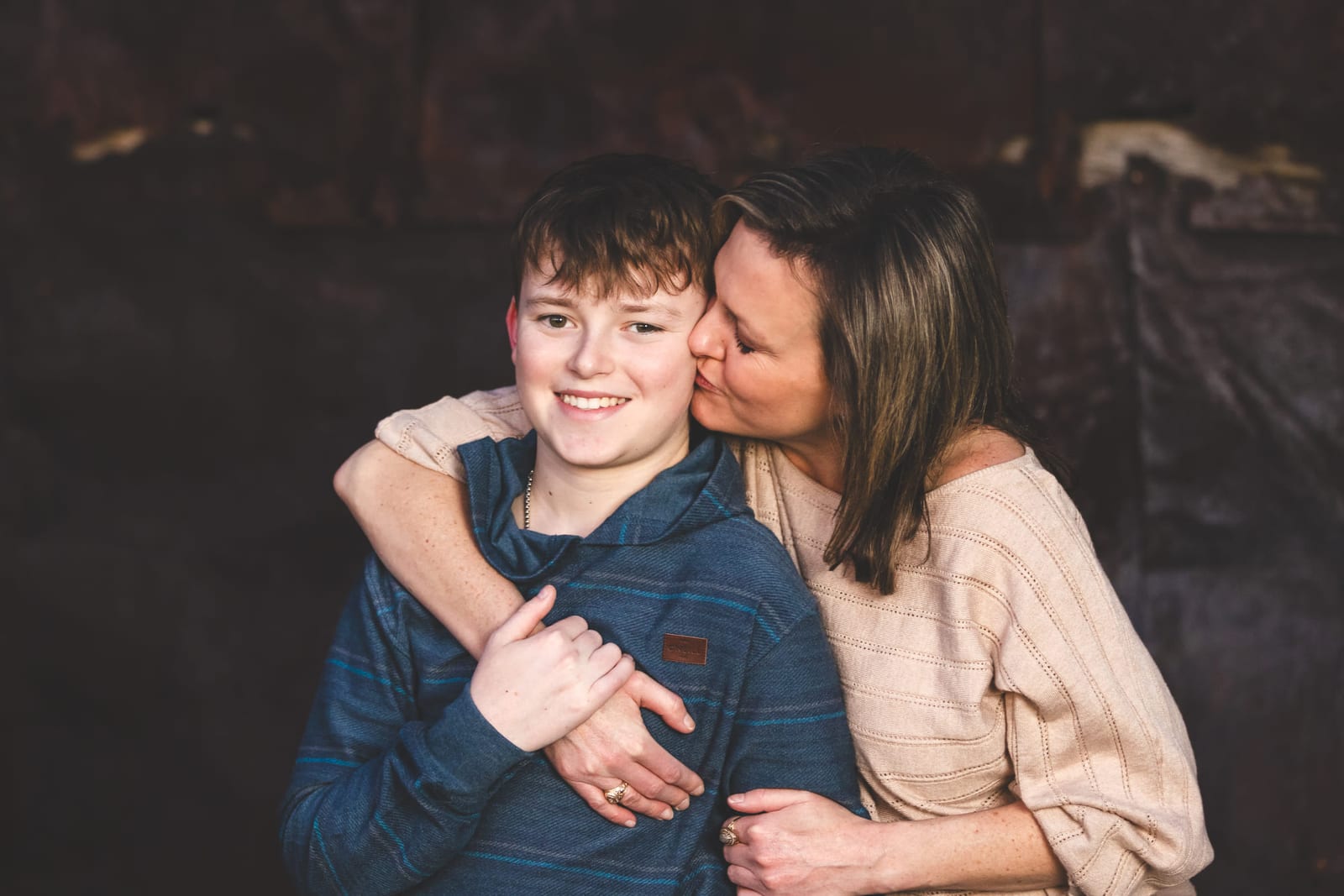 A mother kissing her son’s temple against a dark painted backdrop, both arms wrapped around him.