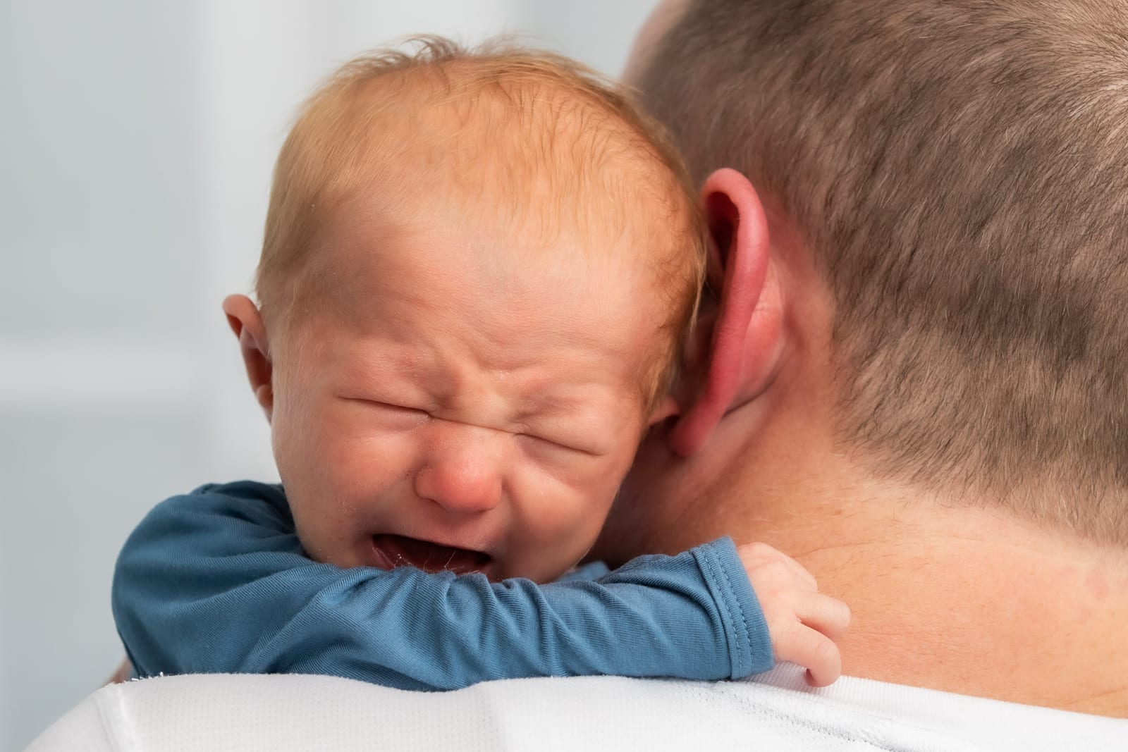 A newborn crying on a father’s shoulder, one small hand curled against his collar.