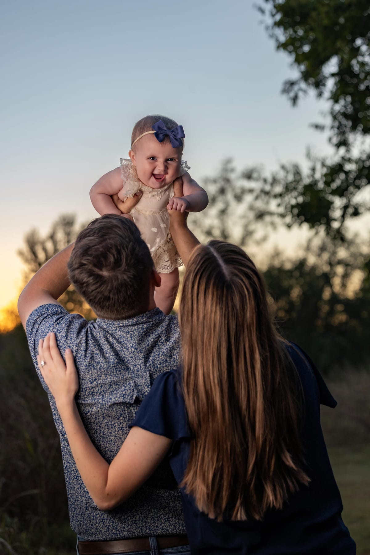 Parents lifting a baby overhead between them in a field at golden hour, seen from behind.