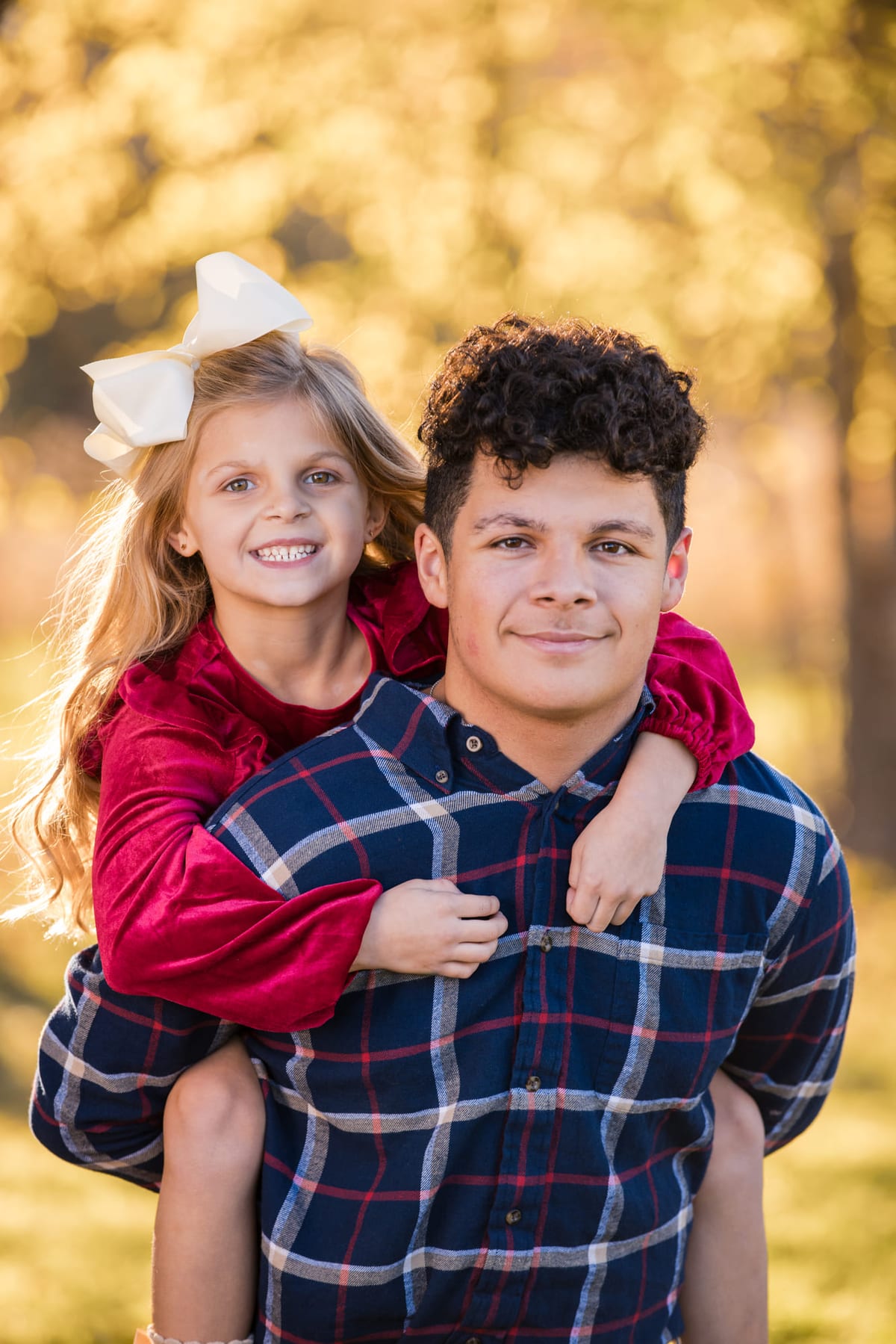 A girl riding on her older brother’s back in plaid, both laughing in a dry field.