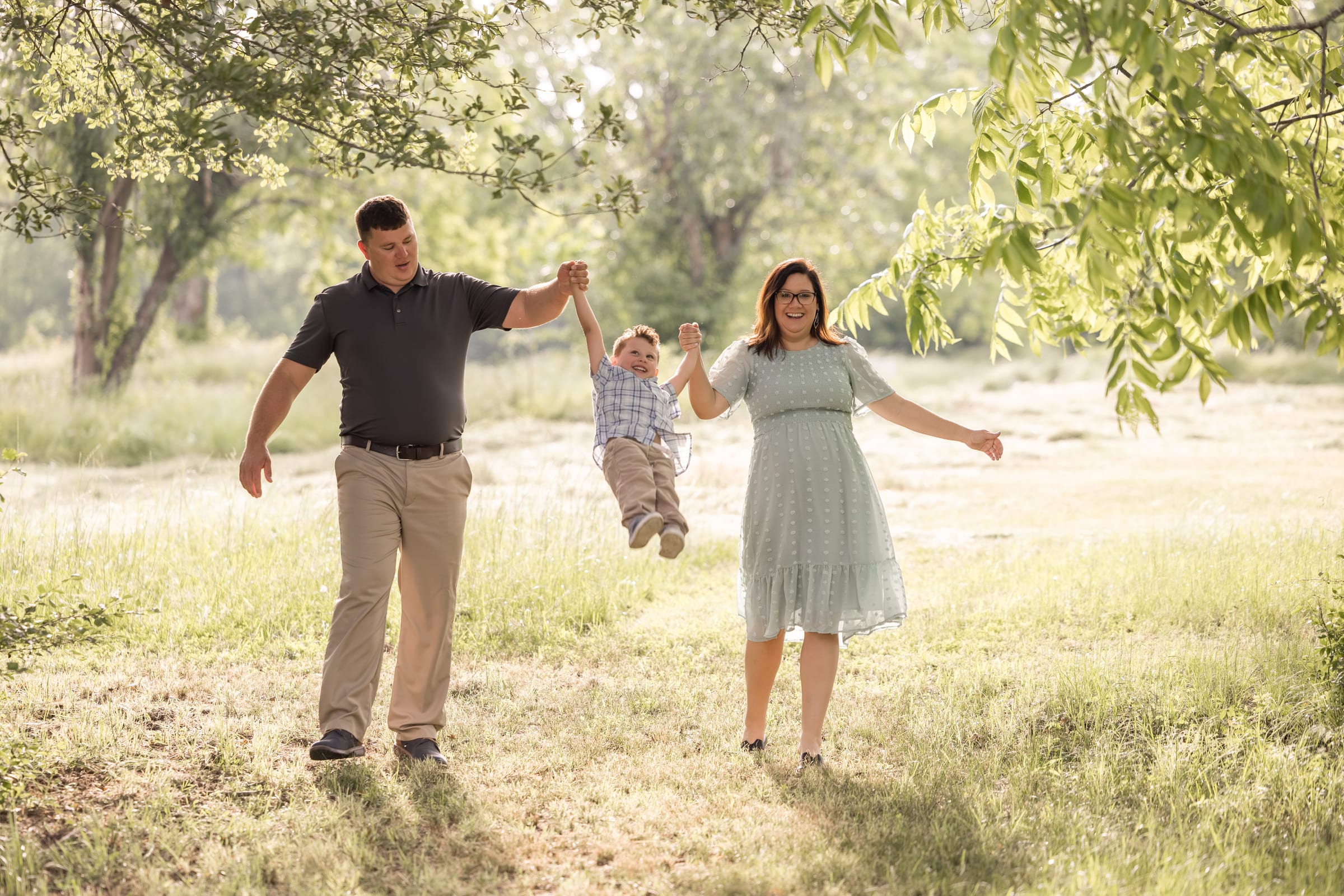 Two parents swinging a small child by the arms between them in a sunlit field, both mid-stride.
