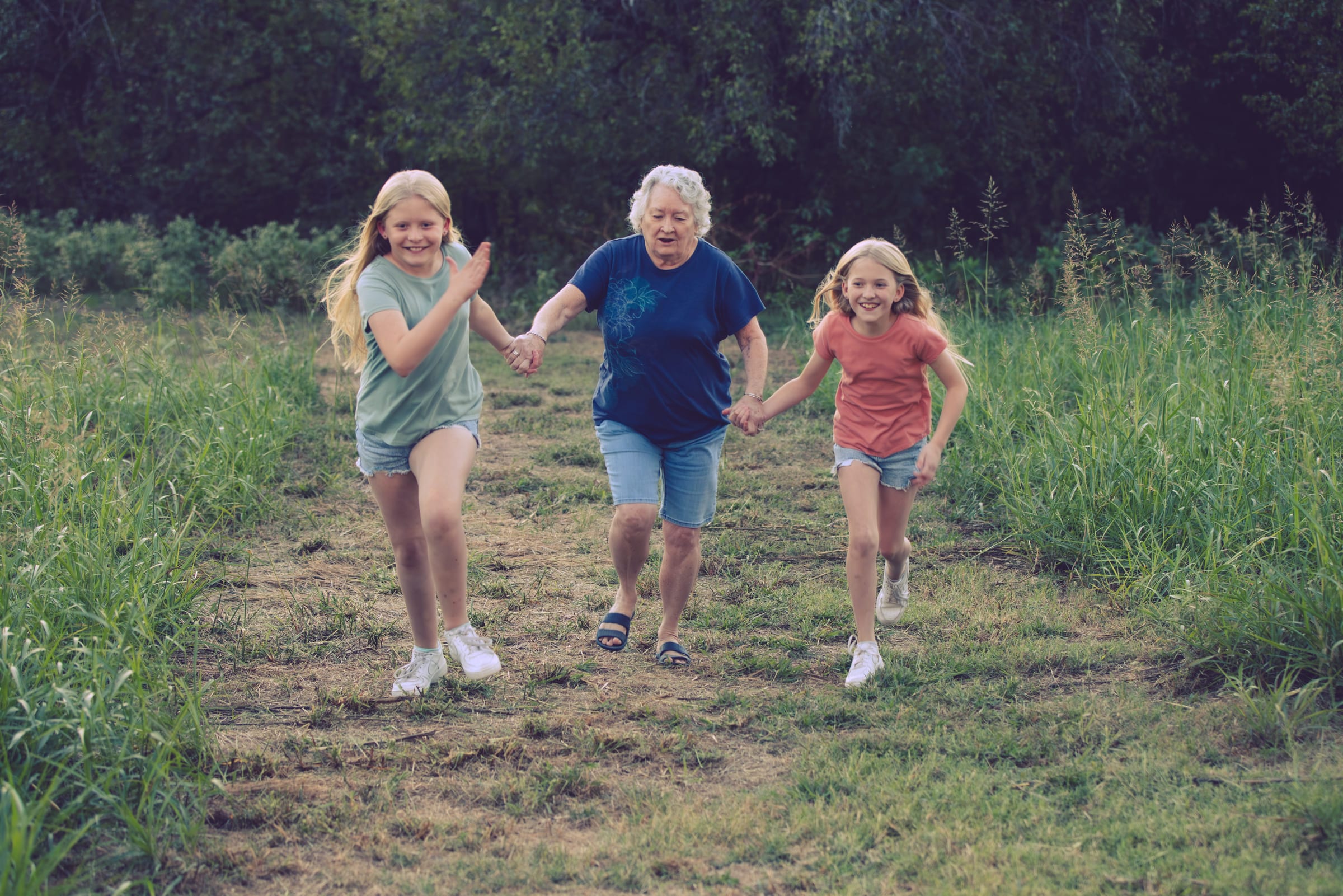 A grandmother running down a grass path holding hands with two young girls, all three laughing.