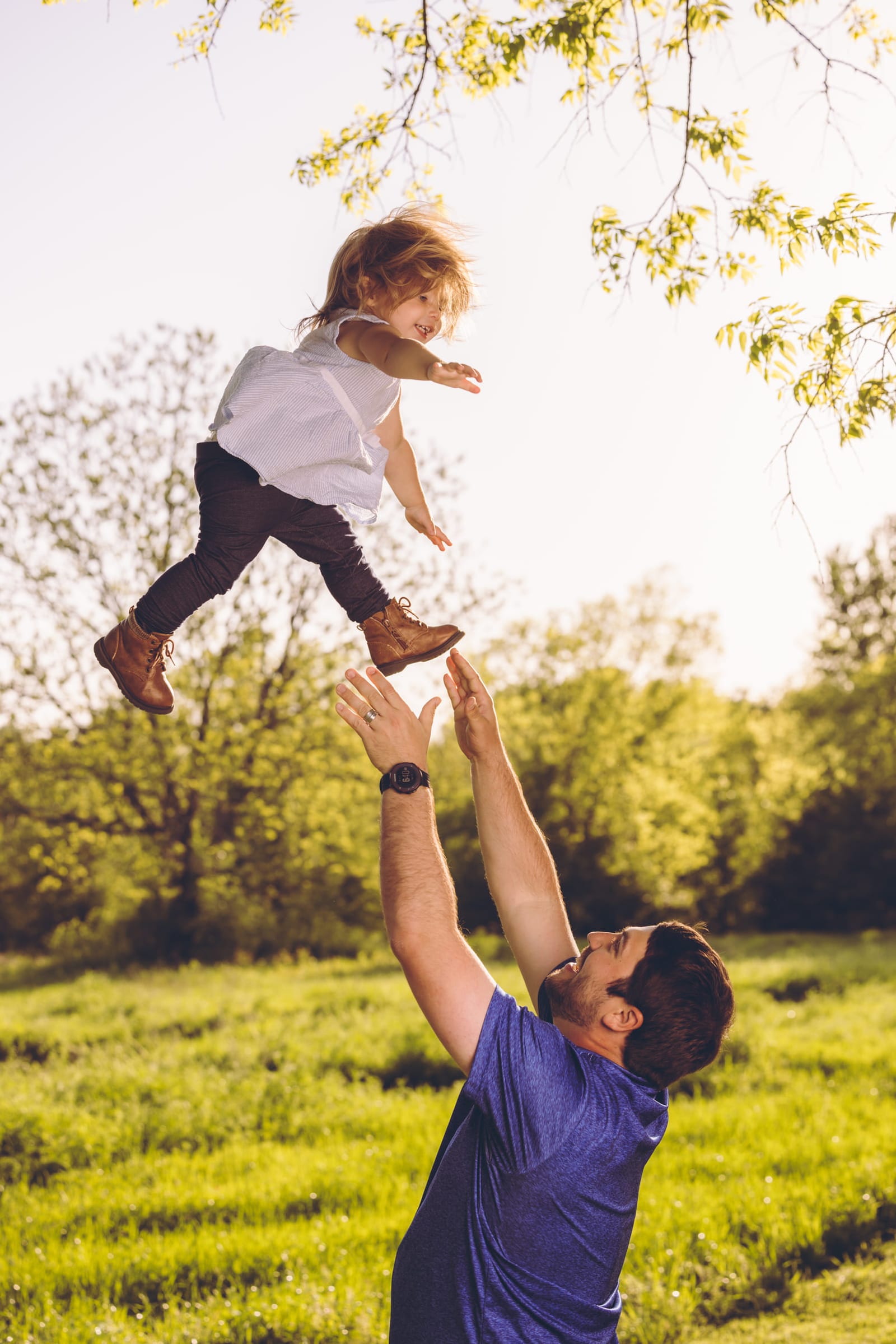 A small child caught in mid-air above her father’s hands, backlit by low sun through the trees.