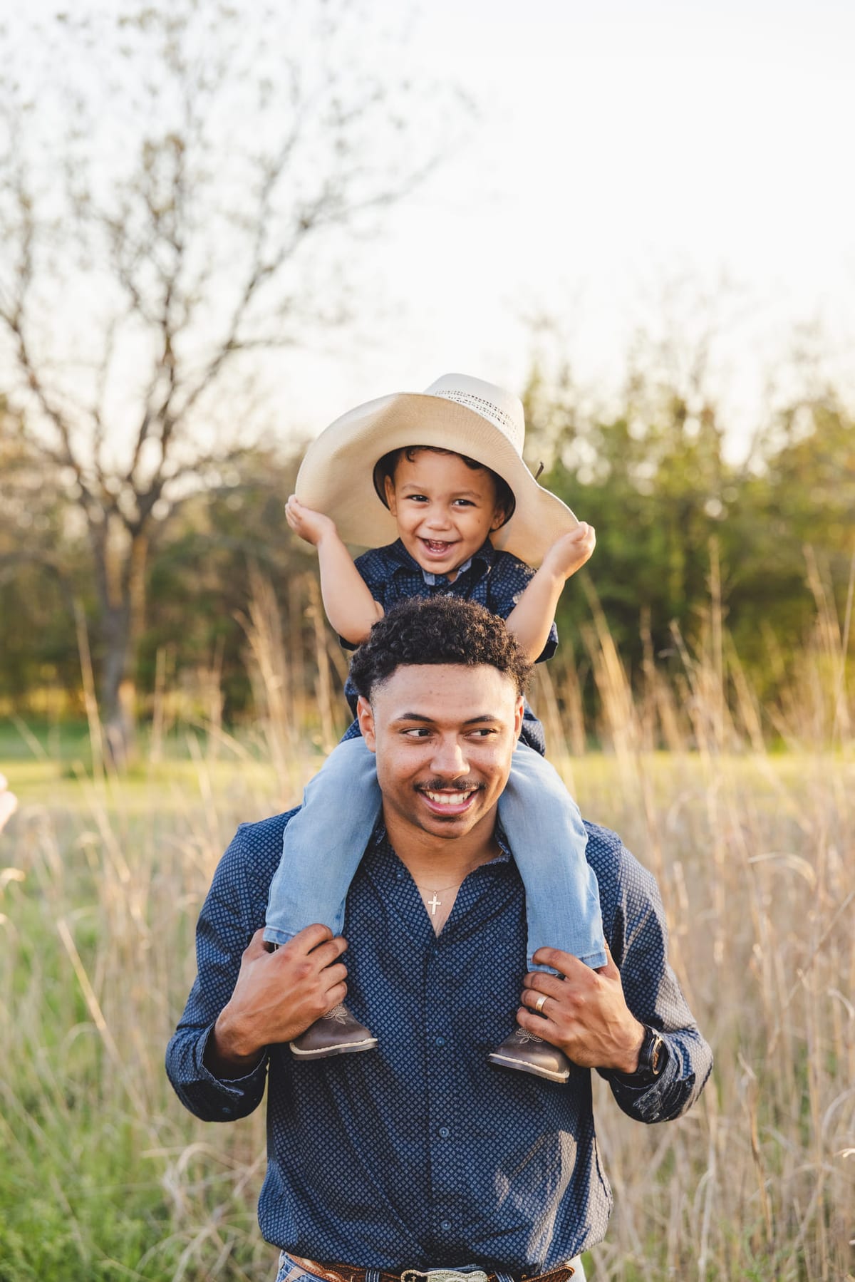 A father carrying his son on his shoulders through tall grass, the boy in a straw hat.