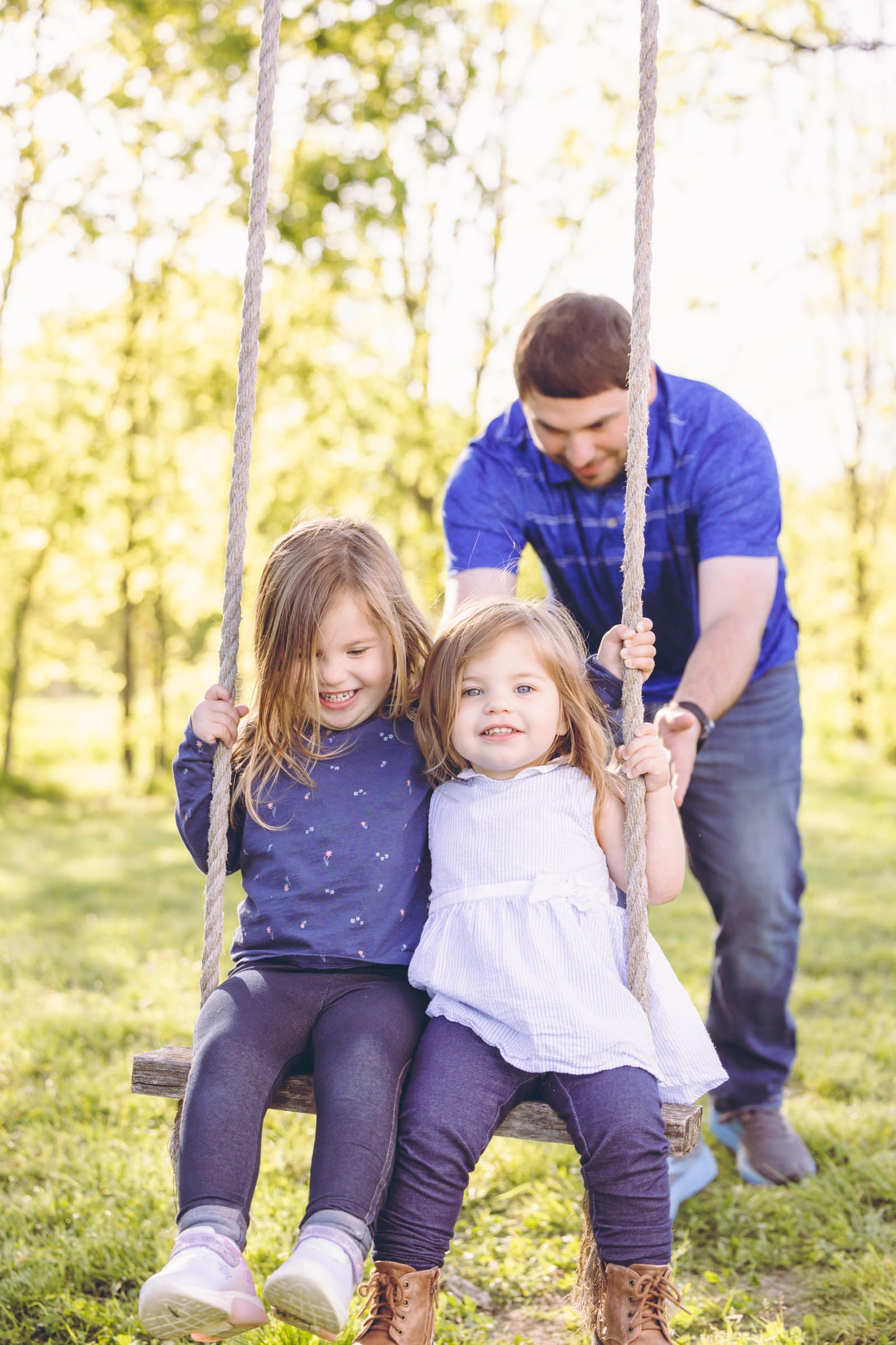 A father pushing his two small daughters on a rope swing in a stand of trees, sunlight coming through behind them.