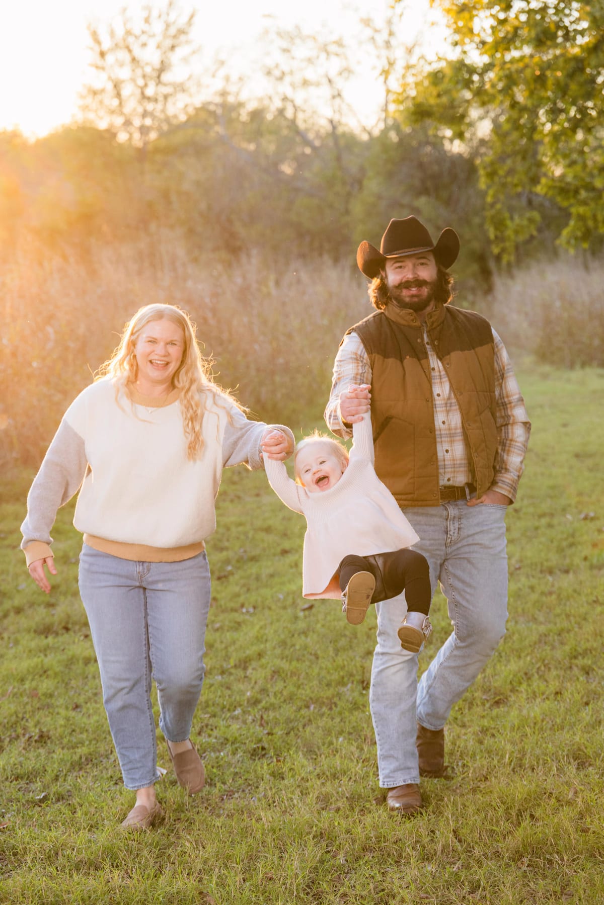Two parents swinging a toddler between them by the hands, barefoot in a winter field.
