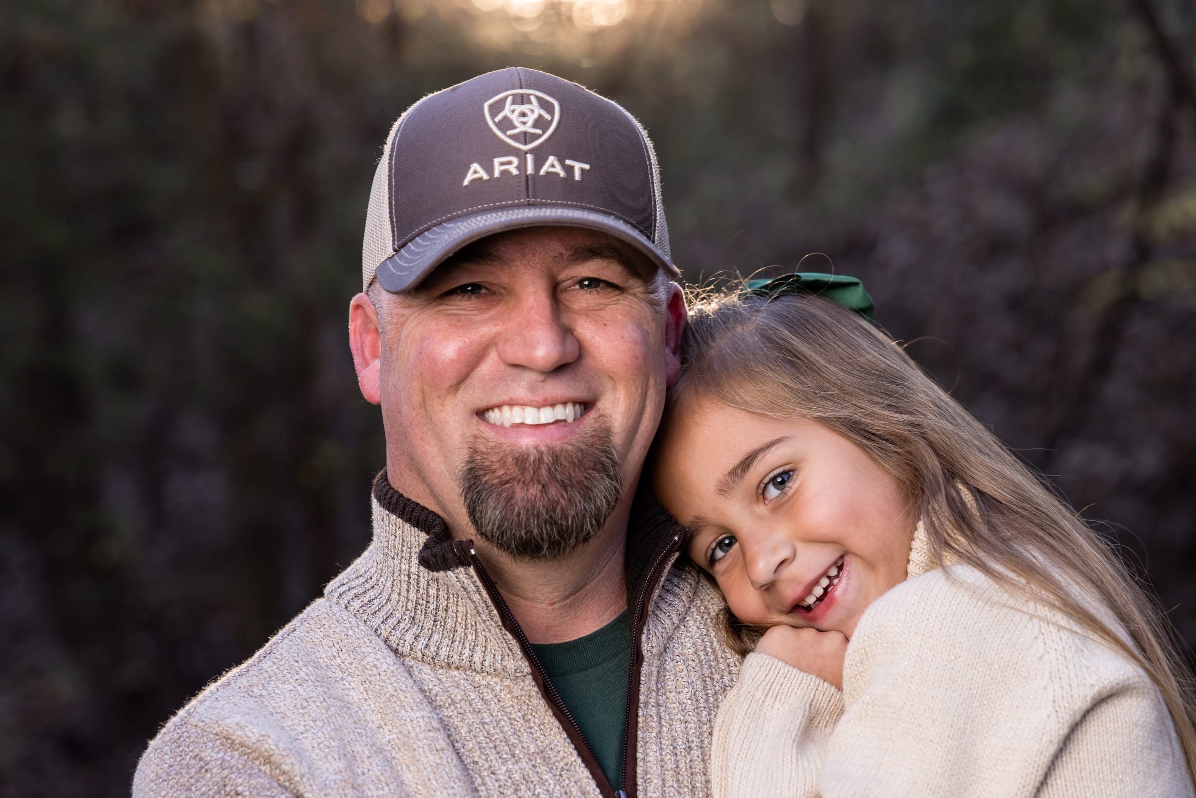 A father in a ball cap cheek to cheek with his young daughter, both grinning at the camera in low evening light.