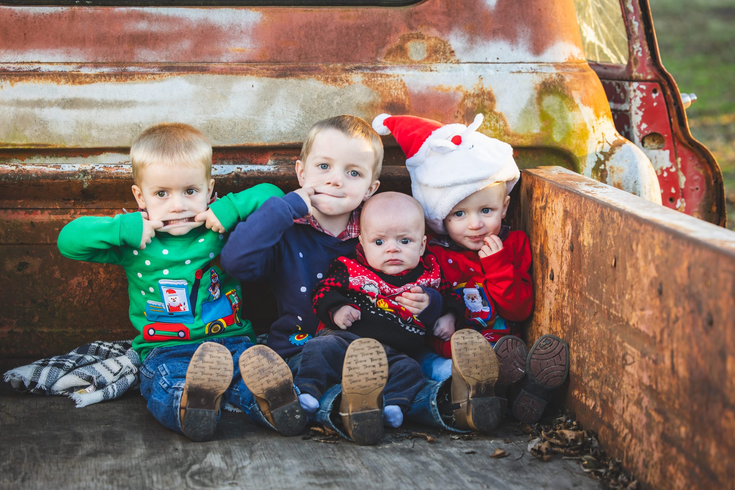 Four small boys in Christmas jumpers lined up along the rusted bed of an old truck, the smallest in a Santa hat.
