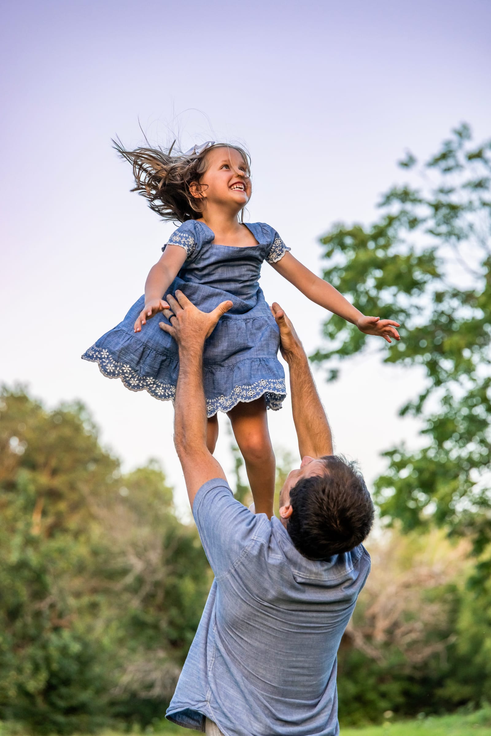 A father throwing his daughter into the air against an open sky, her dress and hair caught mid-flight.