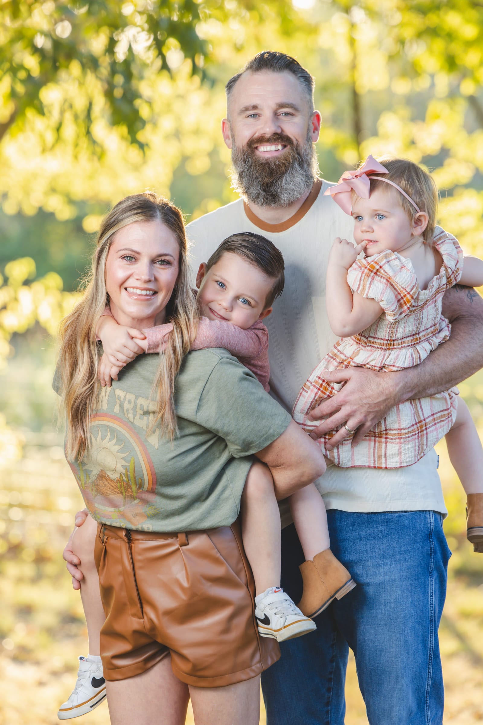 A family of five pressed together in a sunlit clearing, the parents each carrying a child.
