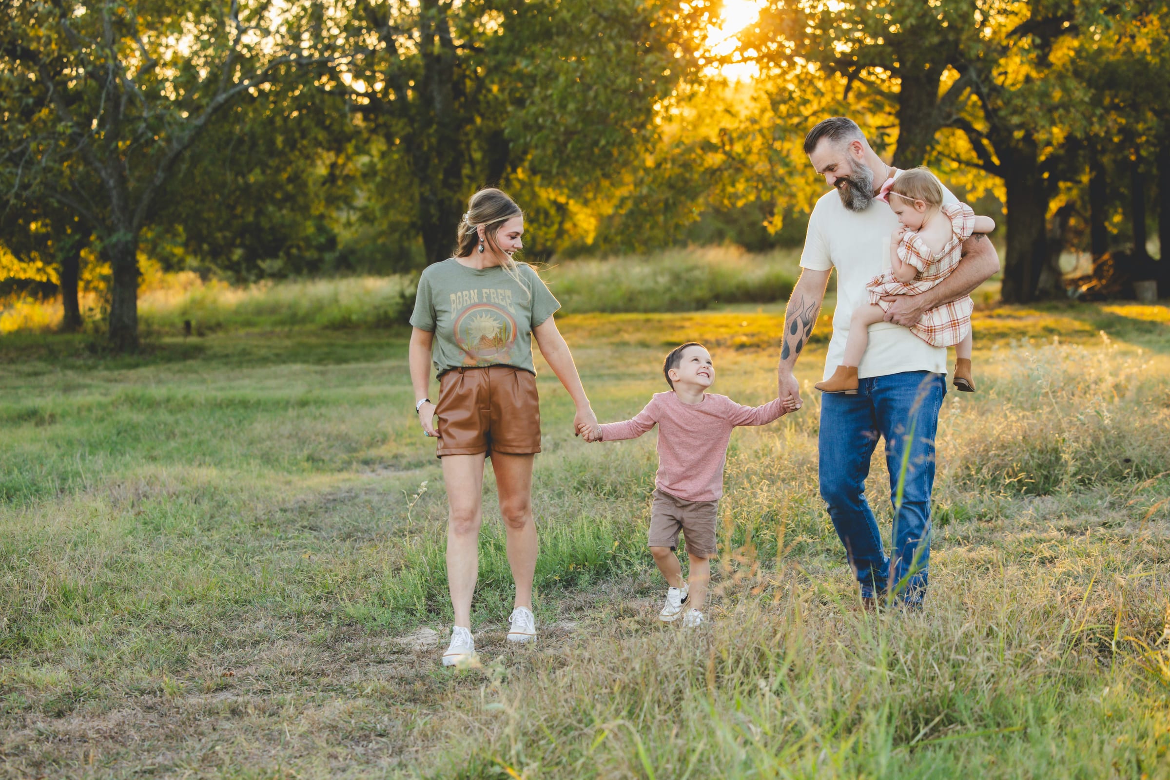 A family of four walking through a sunlit meadow at golden hour, the father carrying a toddler and the mother holding their son’s hand.