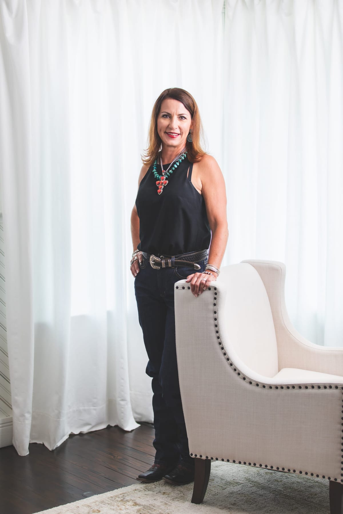 A standing portrait beside an upholstered armchair against a white curtain.