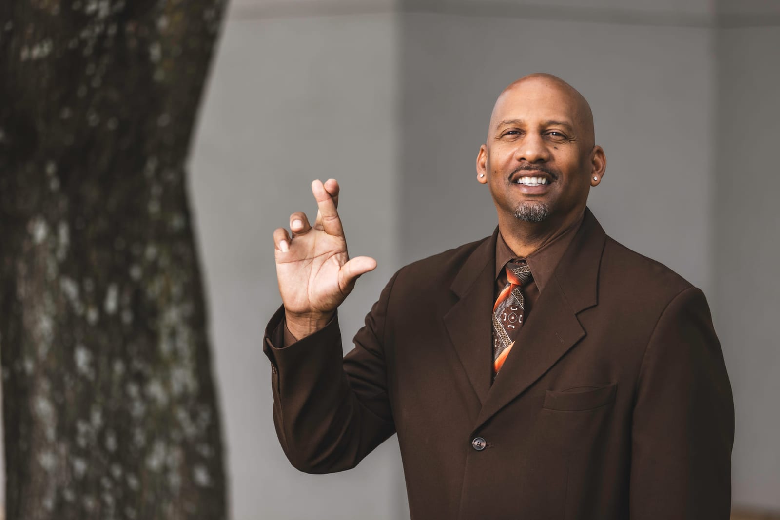 A portrait in a dark suit against a hard-lit grey wall, one hand raised.