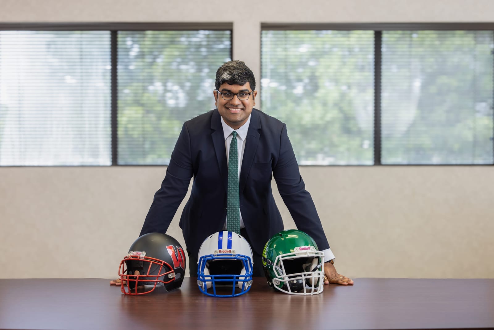 A coach leaning on a table set with three football helmets, windows behind him.