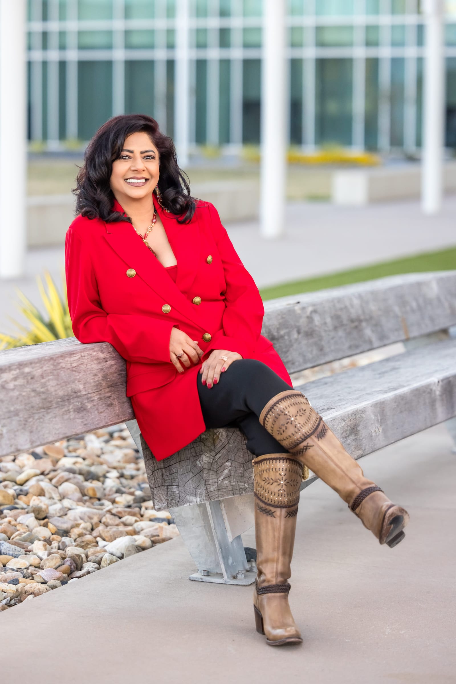 Environmental business portrait of a woman in a red blazer seated on a low wall outside a glass-fronted building.