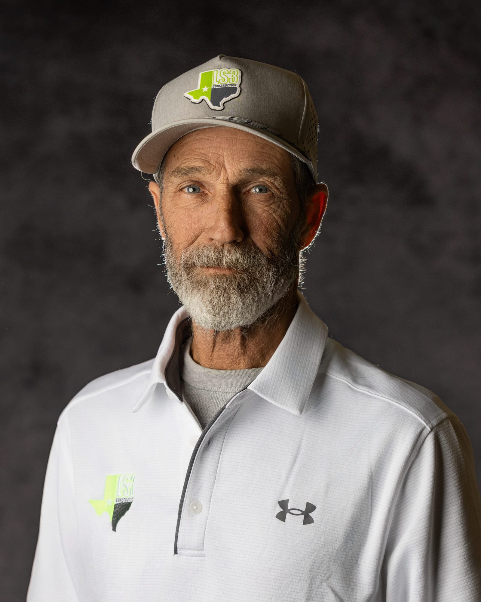 Studio portrait of a bearded man in a branded cap and white polo on a dark mottled ground.