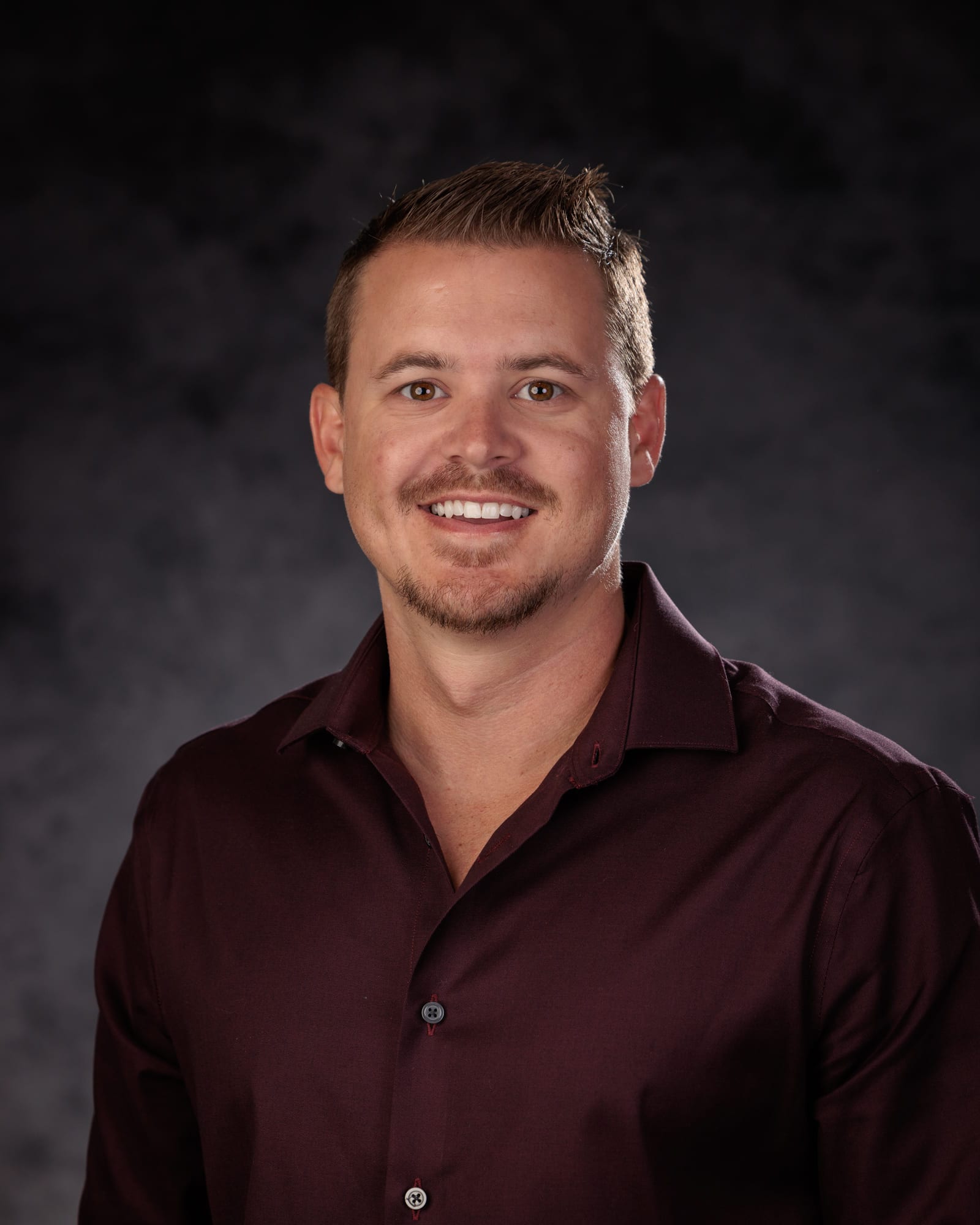 A studio headshot in a maroon shirt against smoke-lit grey.
