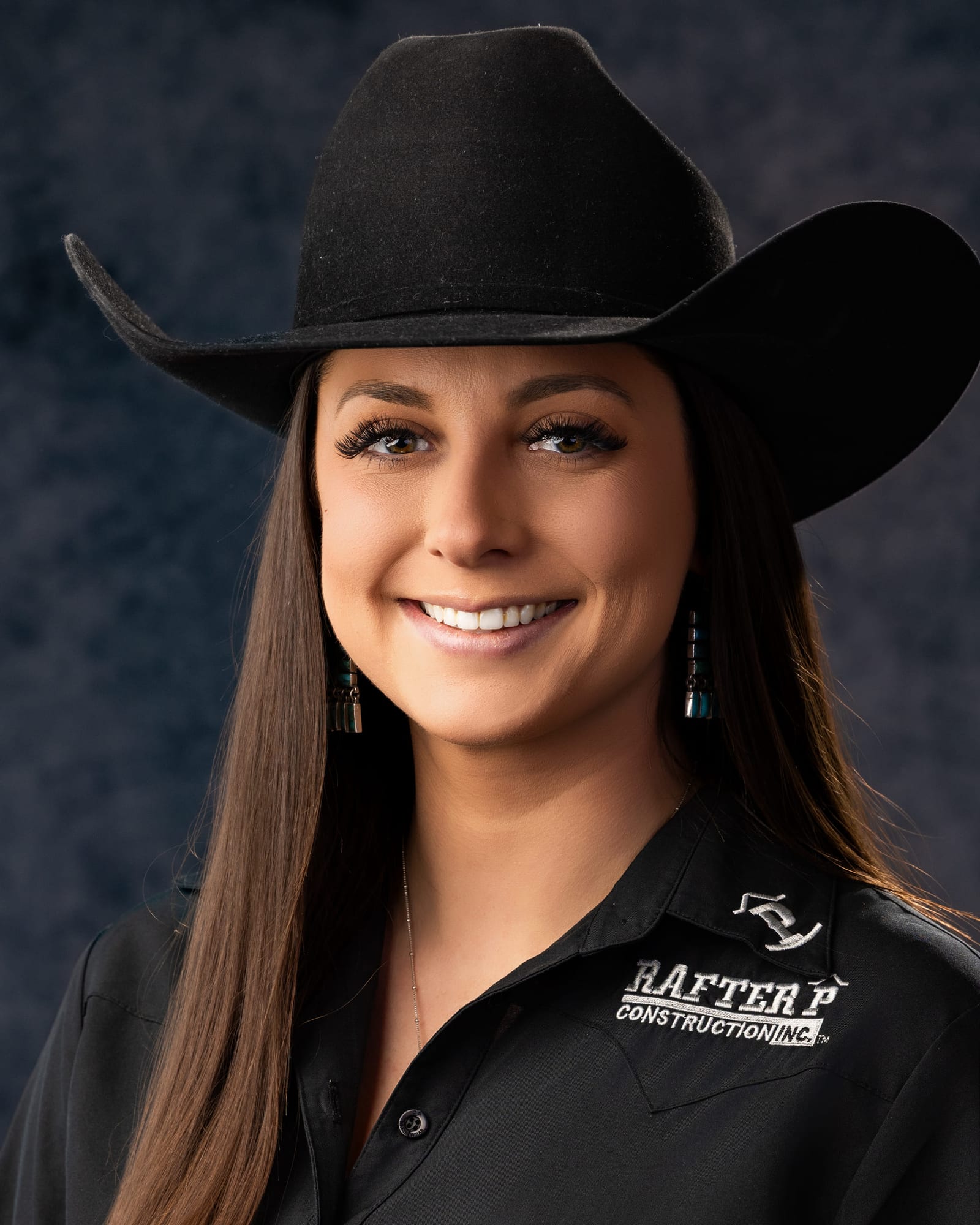 Studio portrait of a woman in a black felt hat and an embroidered work shirt against a mottled grey backdrop.