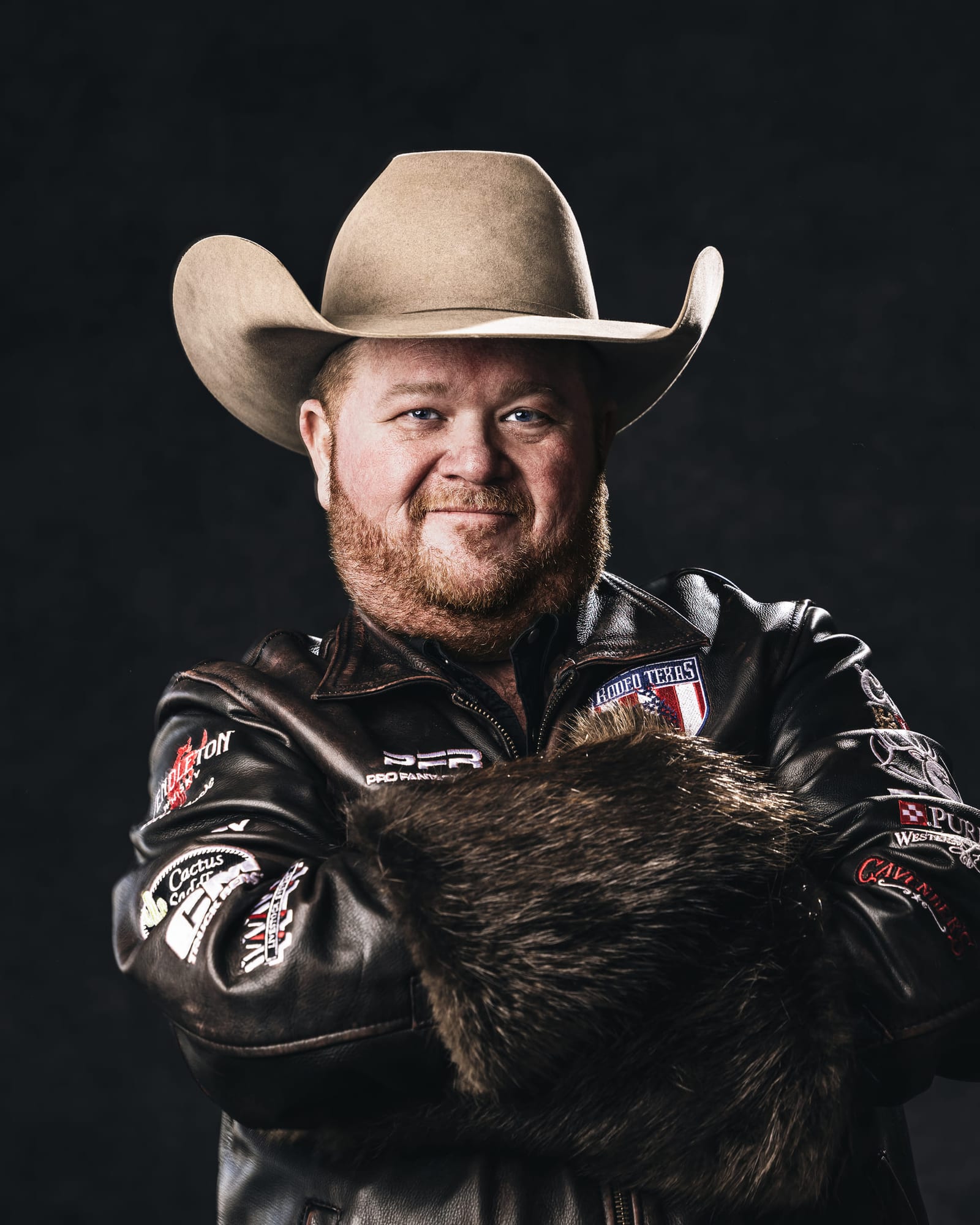 Studio portrait of a man in a straw hat and a fur-collared competition jacket, arms folded, on a black ground.