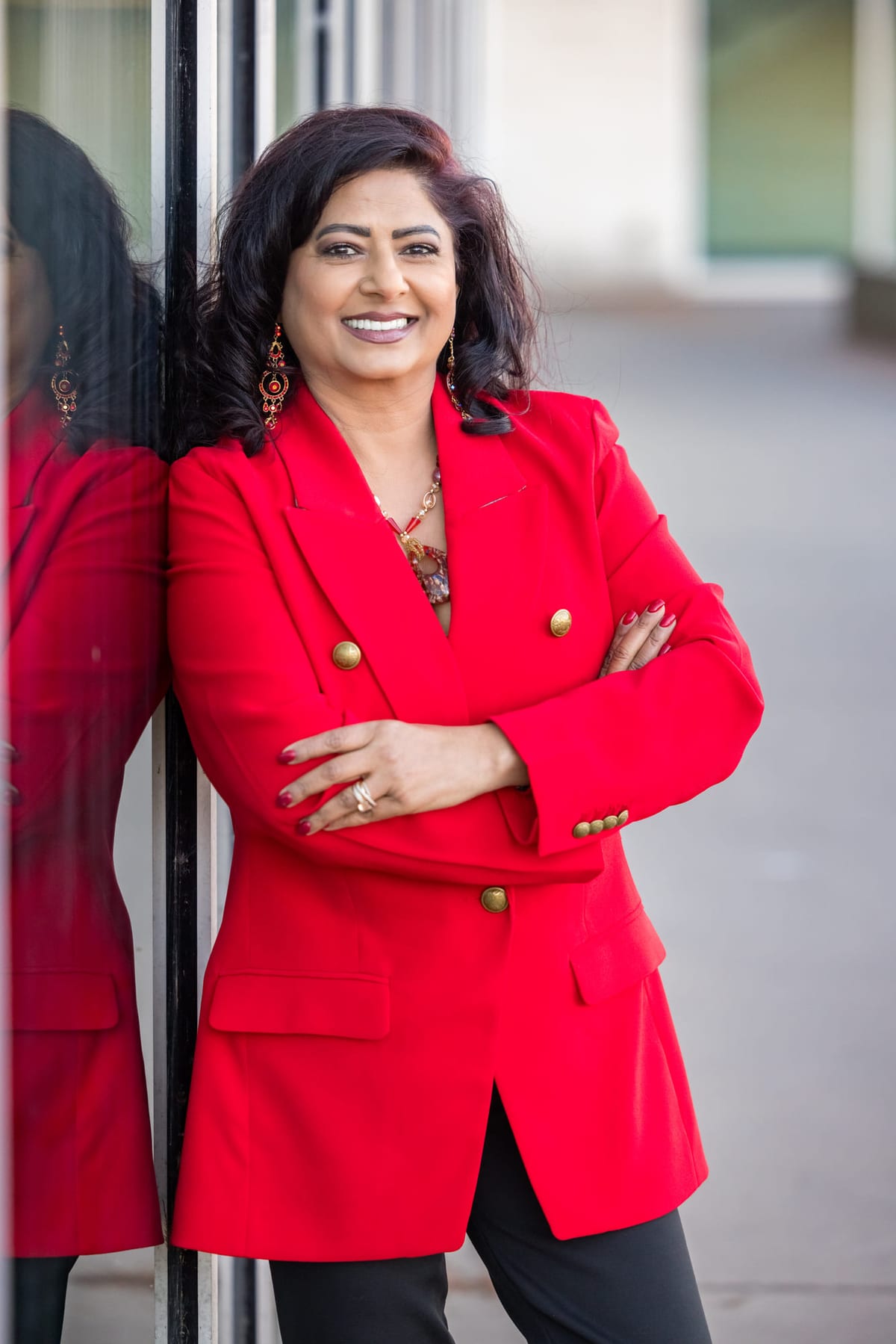 A business portrait in a red blazer against a glass curtain wall, arms folded.