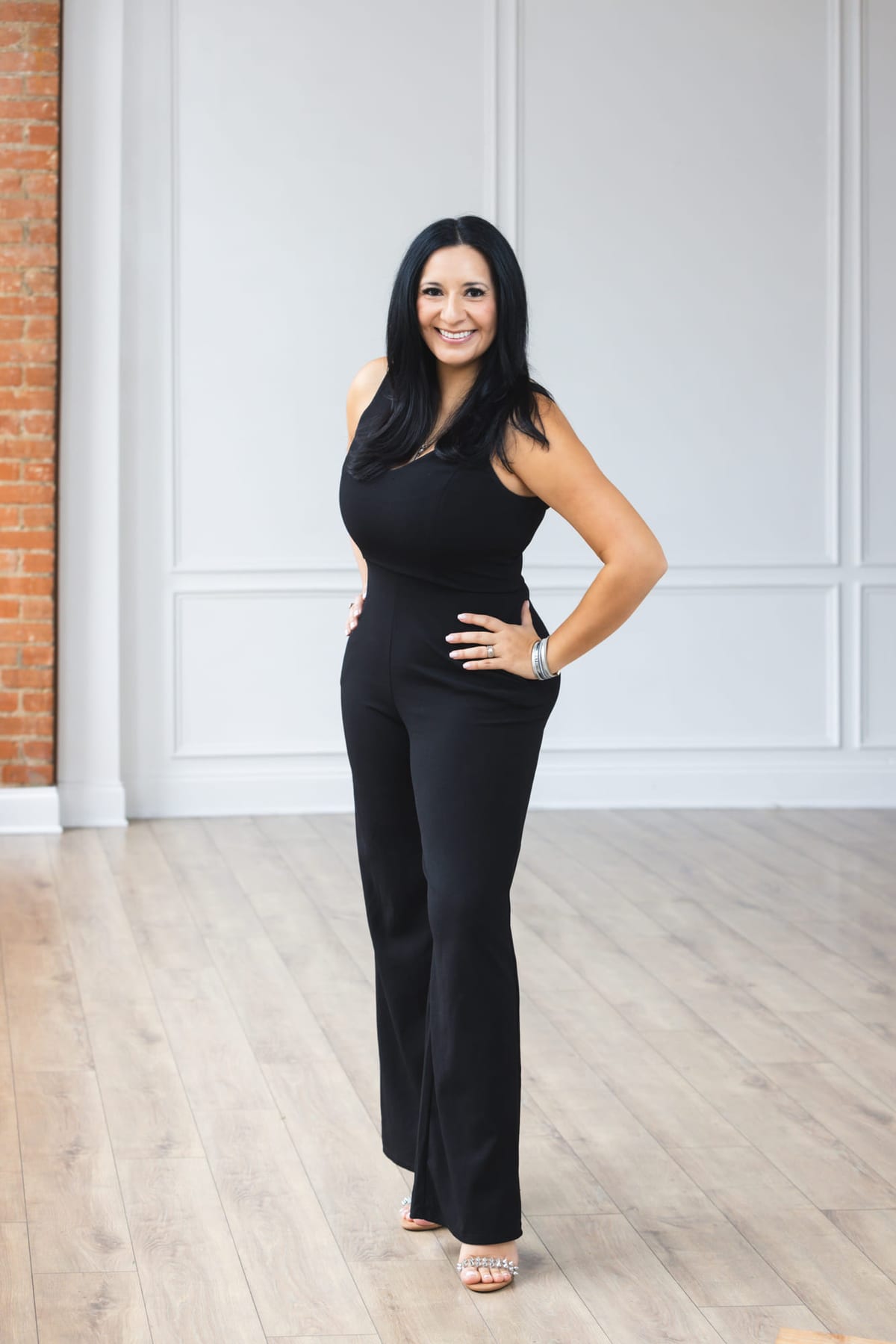 A full-length business portrait in black against a panelled white wall.