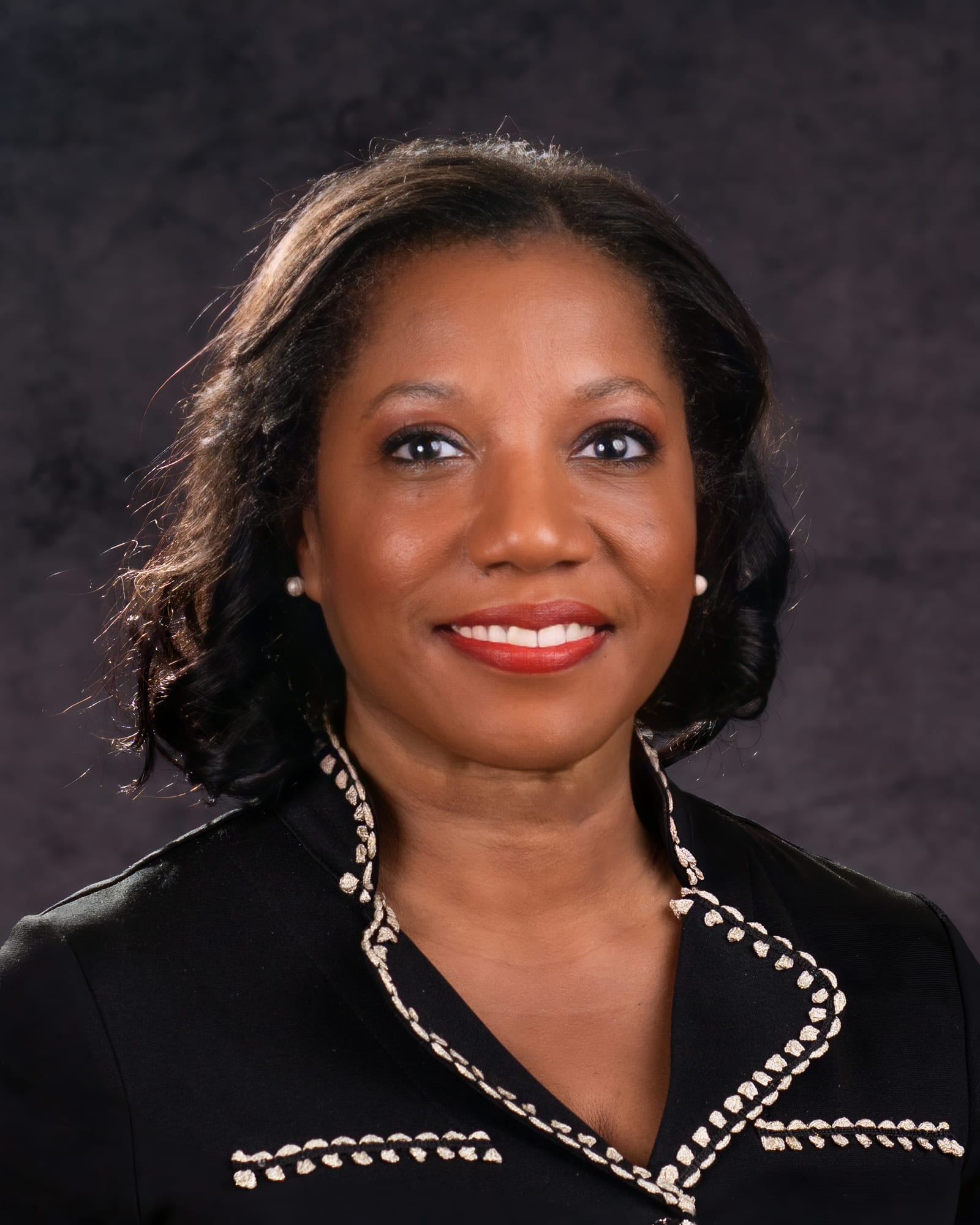 A studio headshot on a plum backdrop, in a trimmed black jacket.