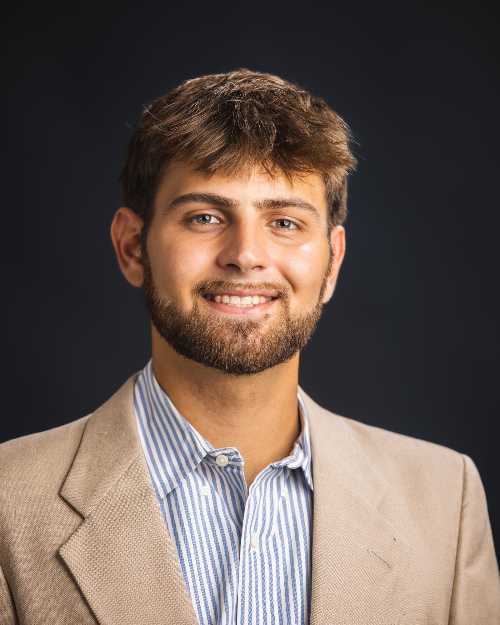 A studio headshot in a tan jacket over a striped shirt.